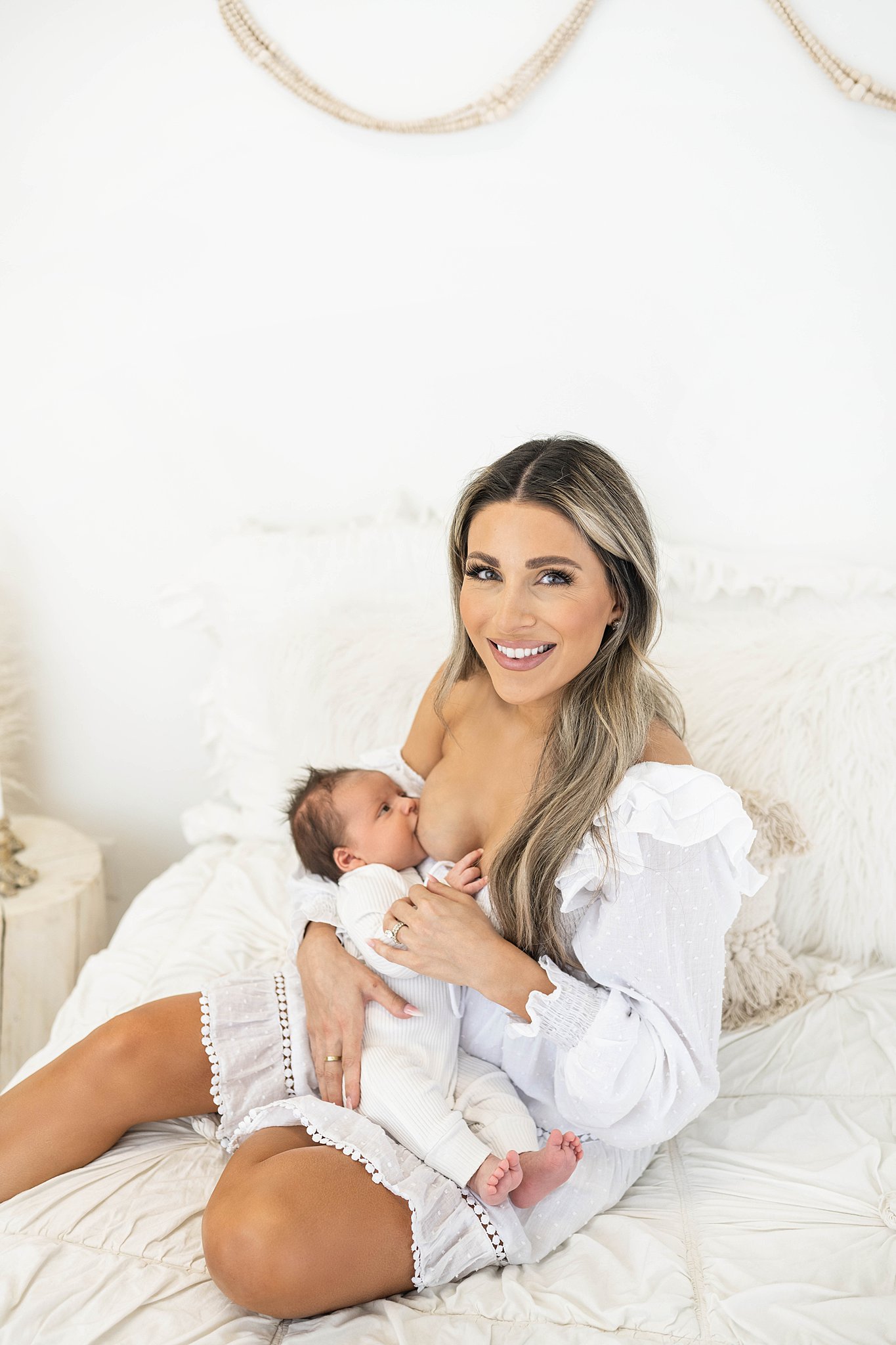 A mother in a white dress sits on a bed while breastfeeding her newborn baby wearing a white onesie Northwest Obstetrics and Gynecology