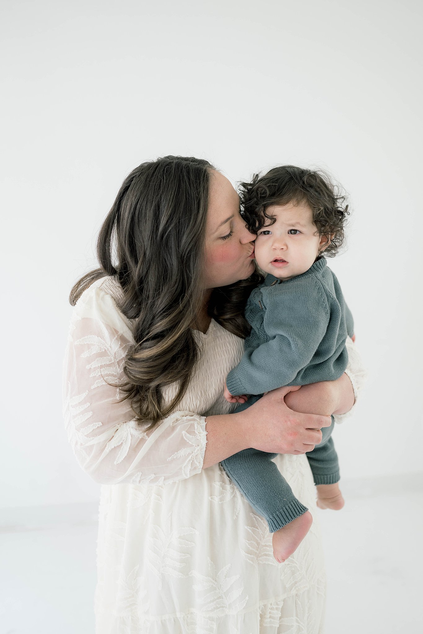 A mother in a white lace dress stands in a studio kissing the cheek of her toddler son with dark curly hair Oklahoma city daycare