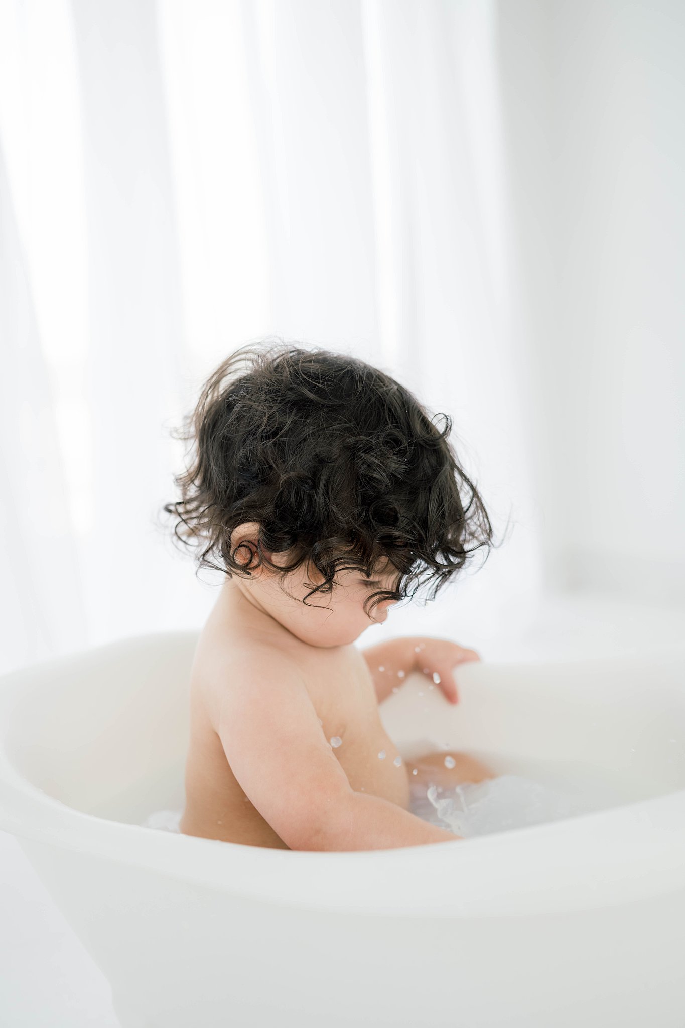 A toddler plays in the water of a small white bathtub in front of a window with white curtains baby furniture Oklahoma