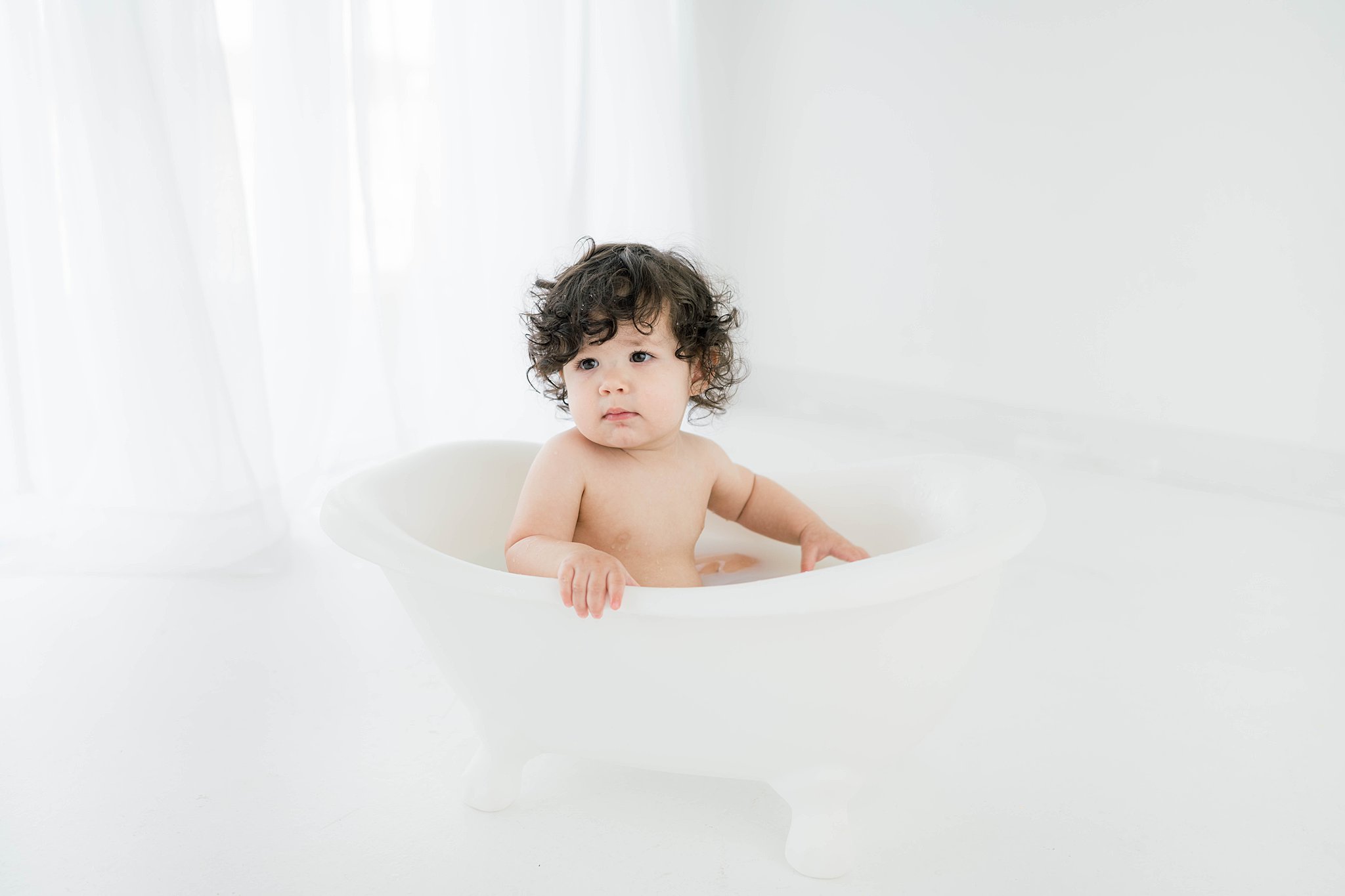 A young toddler with dark curly hair sits on a tiny bathtub in a white room in front of a window baby furniture Oklahoma
