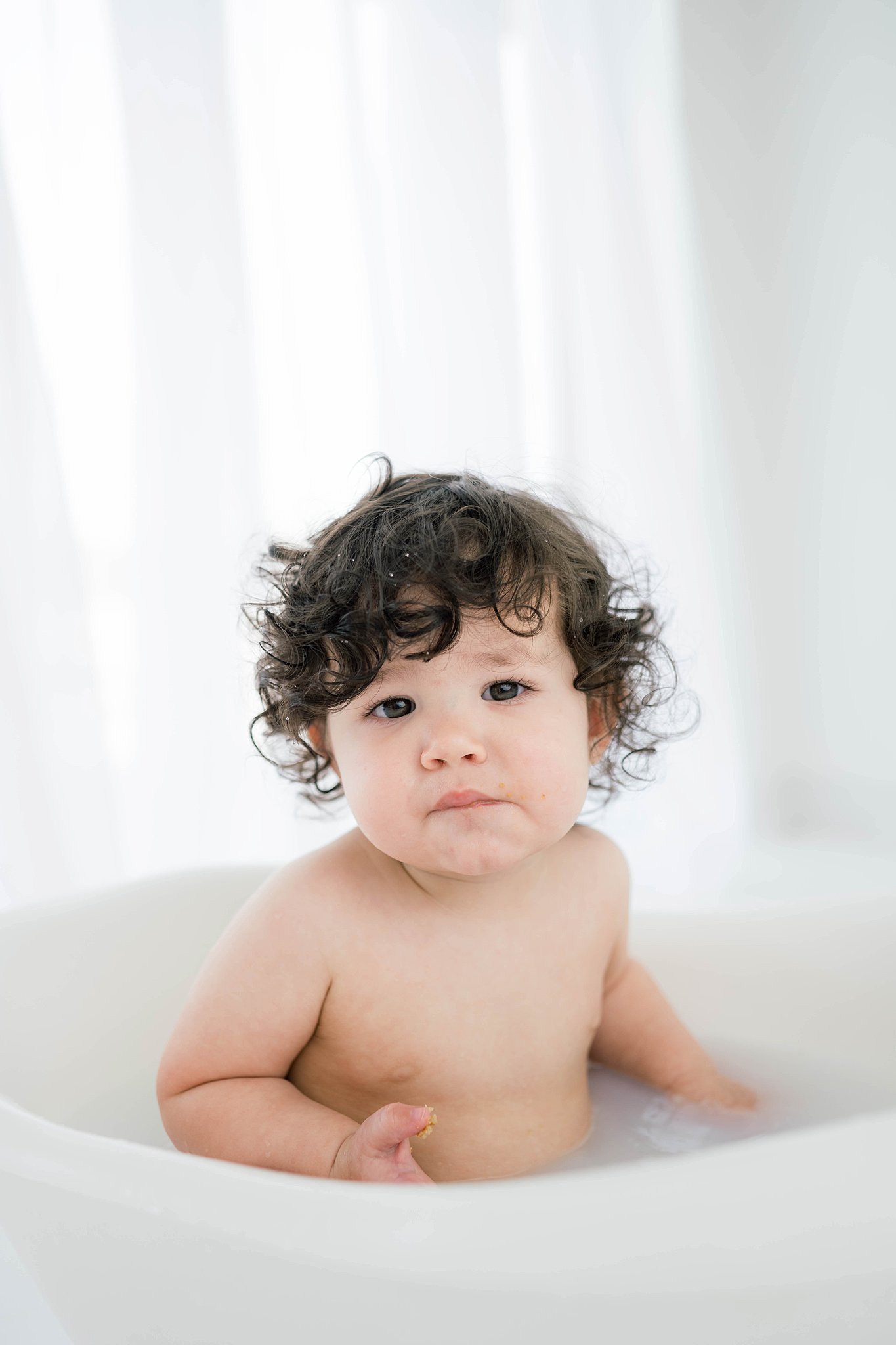 A young toddler with dark curly hair plays in a bathtub in front of a tall window with white curtains baby furniture Oklahoma