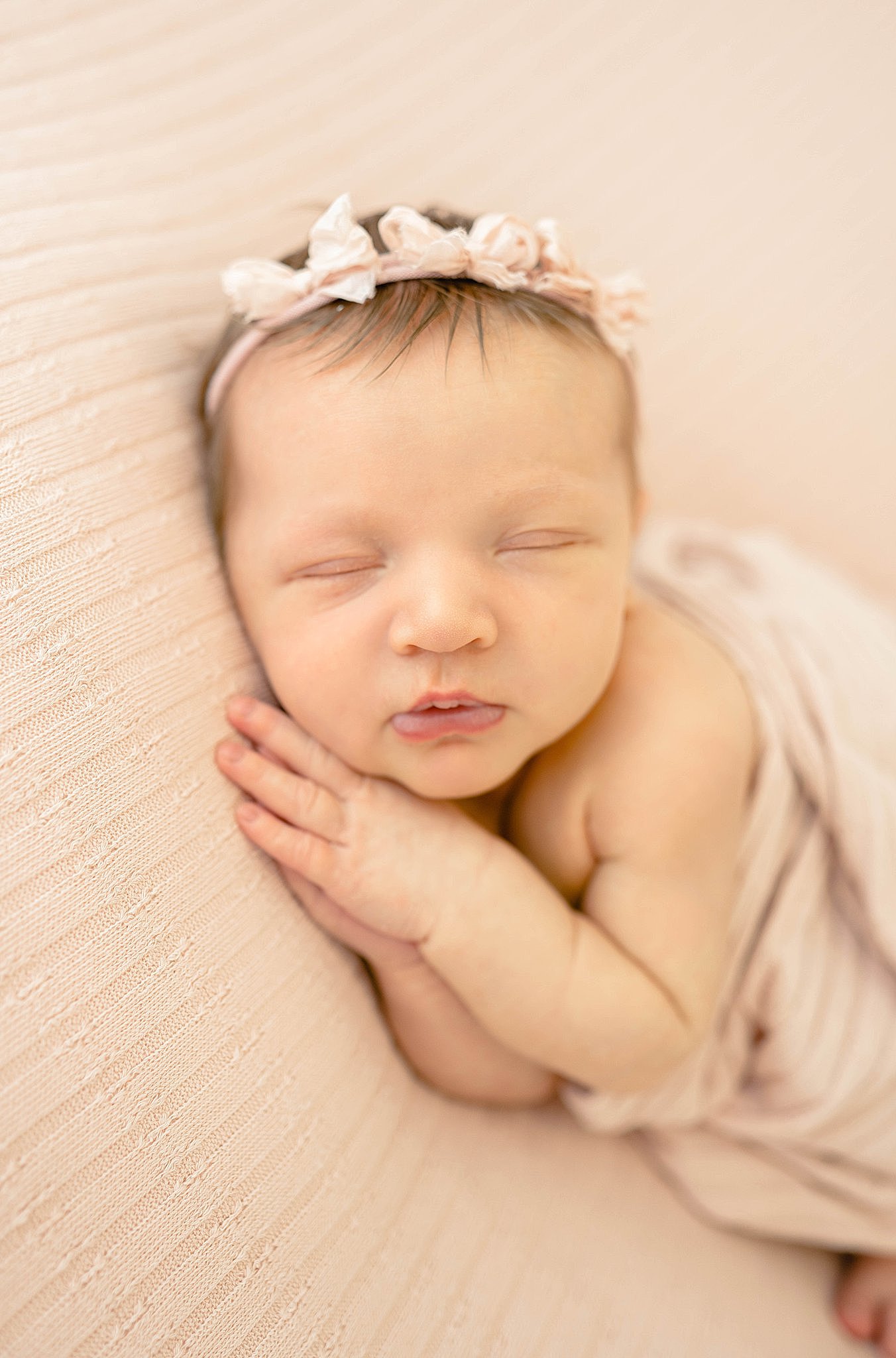 A newborn baby girl sleeps on her hands while wrapped in a pink blanket and wearing a pink bow headband beautifully connected