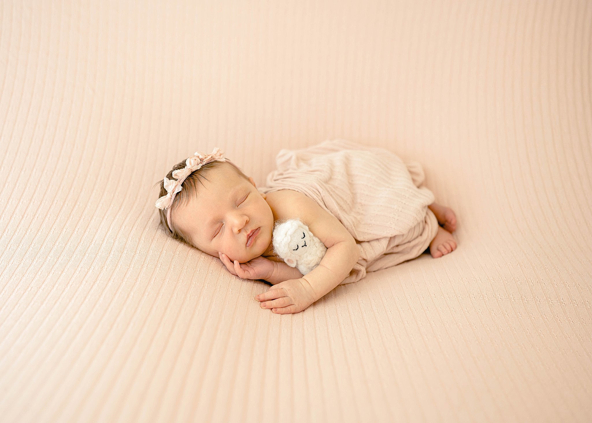 A newborn baby girl snuggles and sleeps with a tiny knit lamb on a pink bed beautifully connected