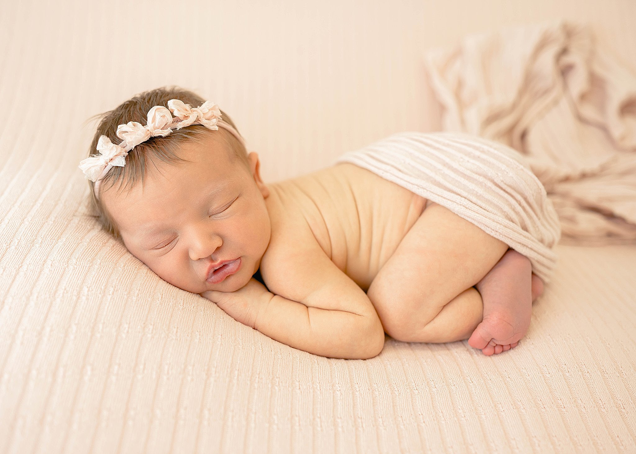 A newborn baby girl sleeps with her face squished on a pillow covered by a pink blanket in froggy pose