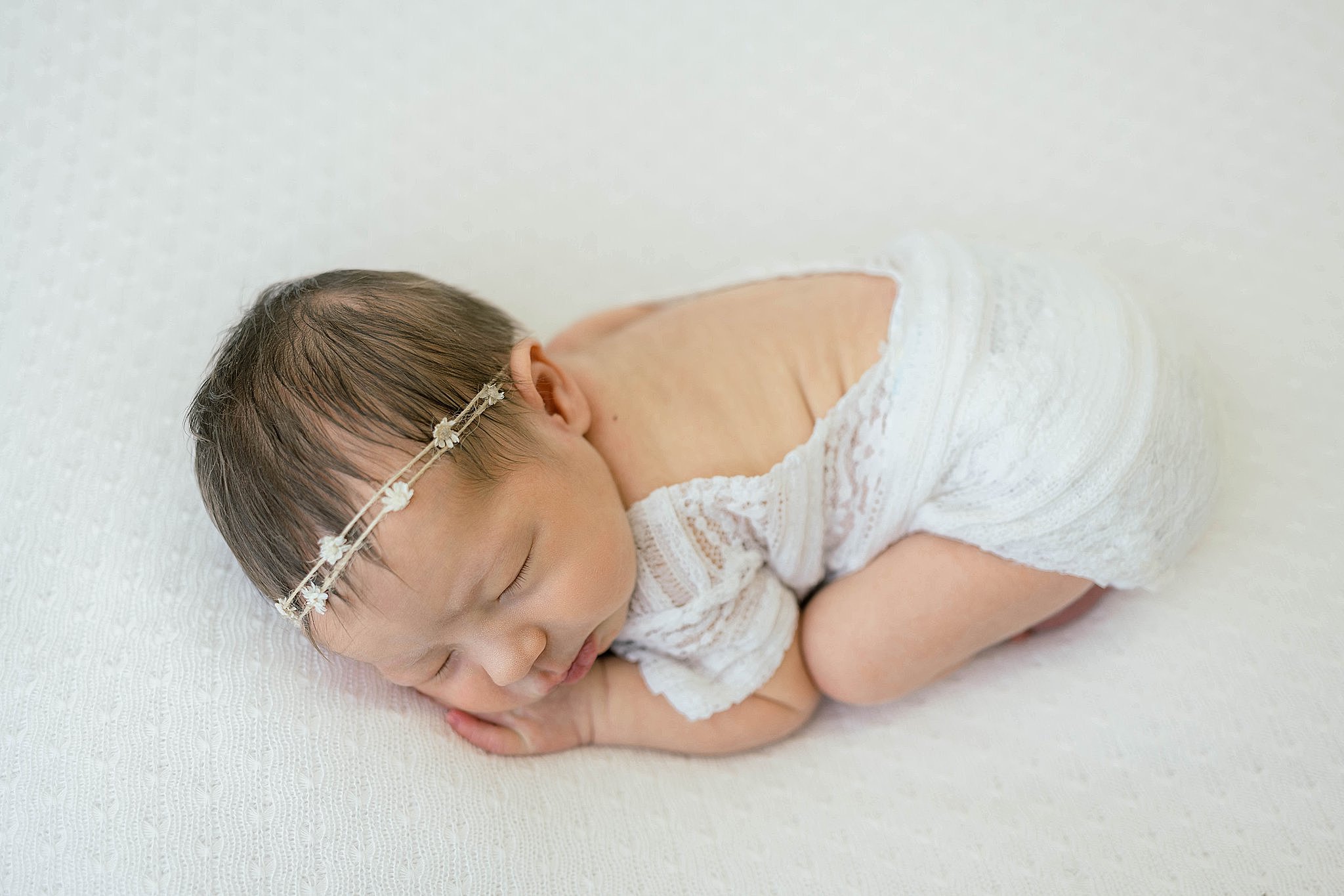 A newborn baby girl sleeps in froggy pose in a white lace onesie wearing a string flower headband bella baby