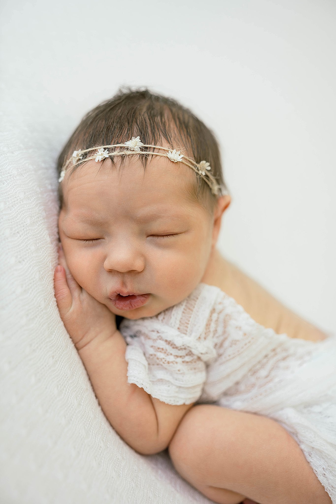 A newborn baby sleeps on her hands in a white lace onesie bella baby