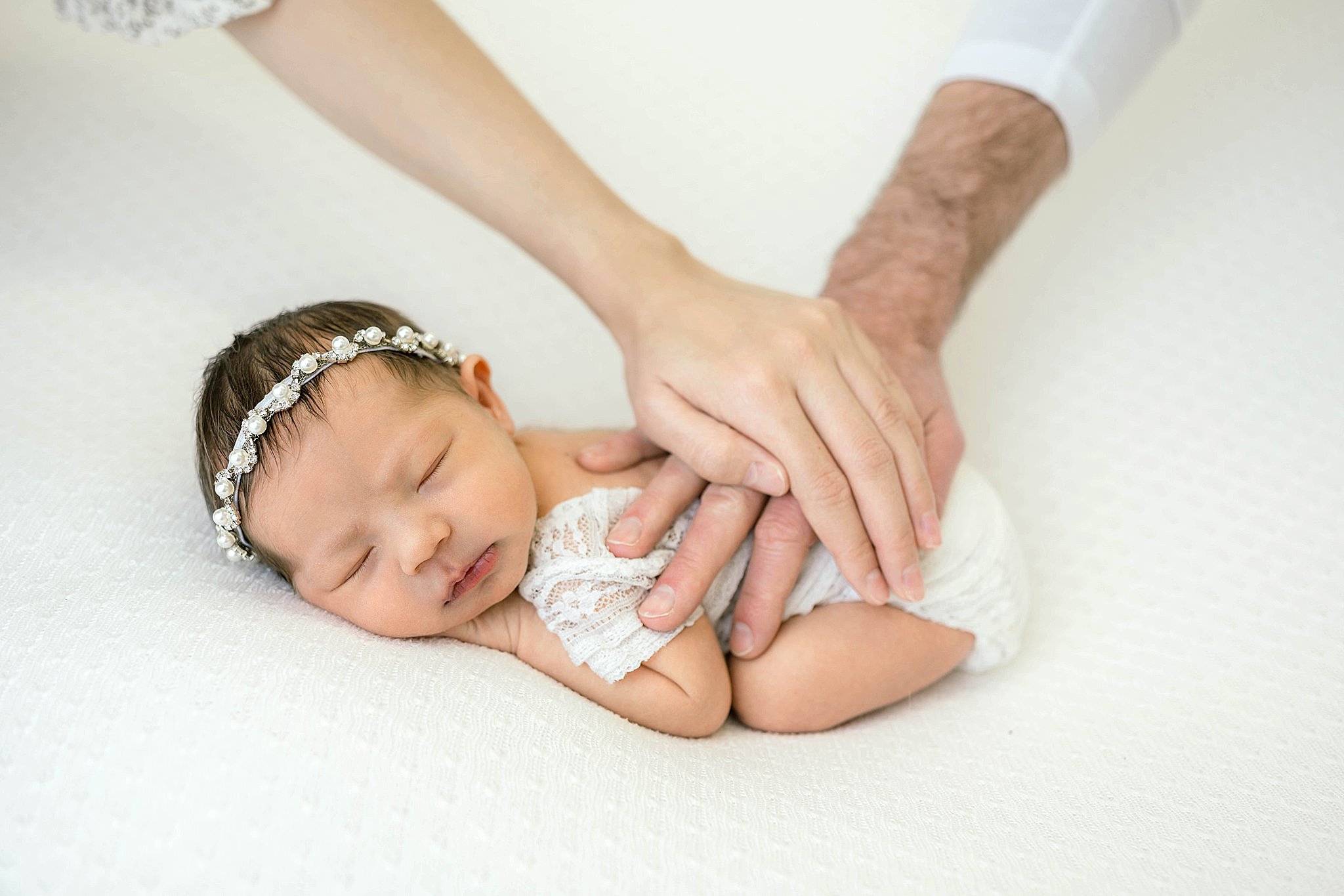 Parent each place a hand on their newborn baby girl while she sleeps on a white bed