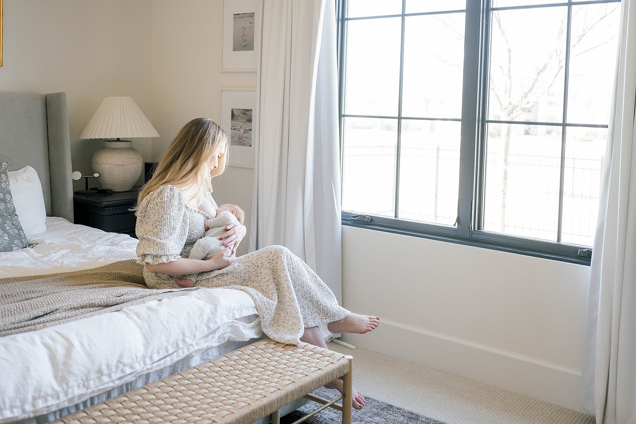 A mother sits on the edge of her bed in front of a large window breastfeeding her newborn baby doula oklahoma city