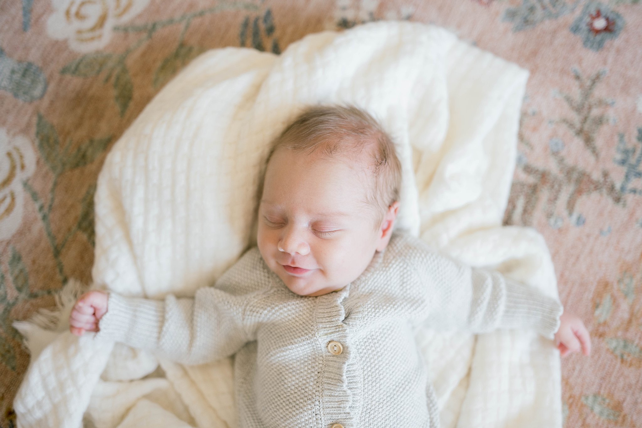 A newborn baby in a white knit sweater sleeps with arms out on a white blanket Lay’s World