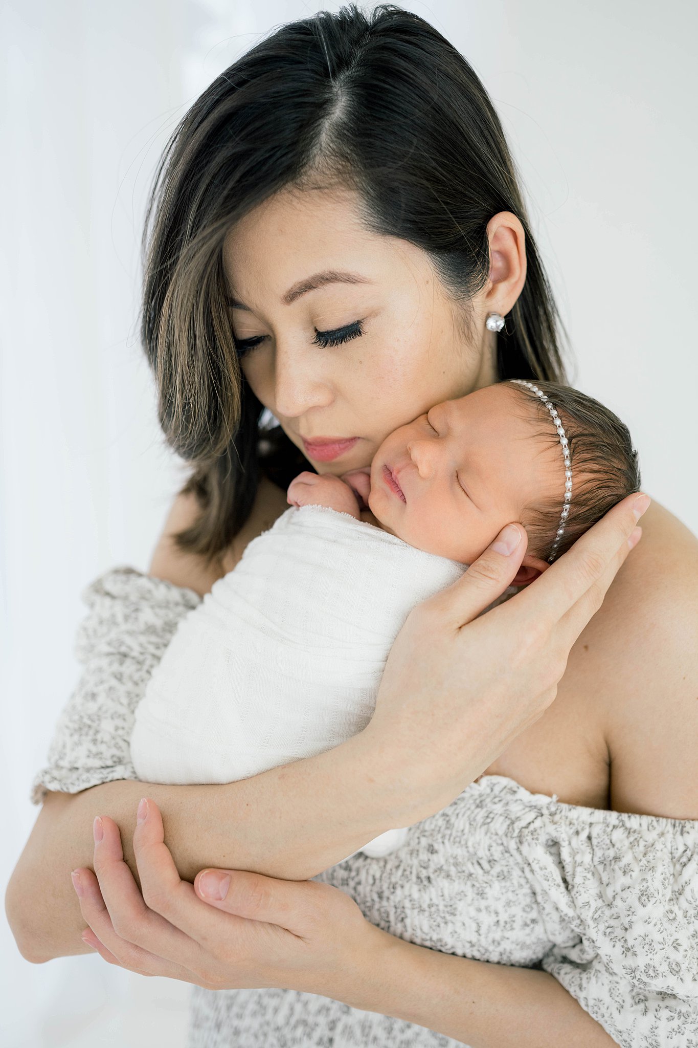 A mother in a pattern off the shoulder dress cuddles her sleeping newborn baby girl on her cheek while standing in a studio maternity clothes oklahoma city