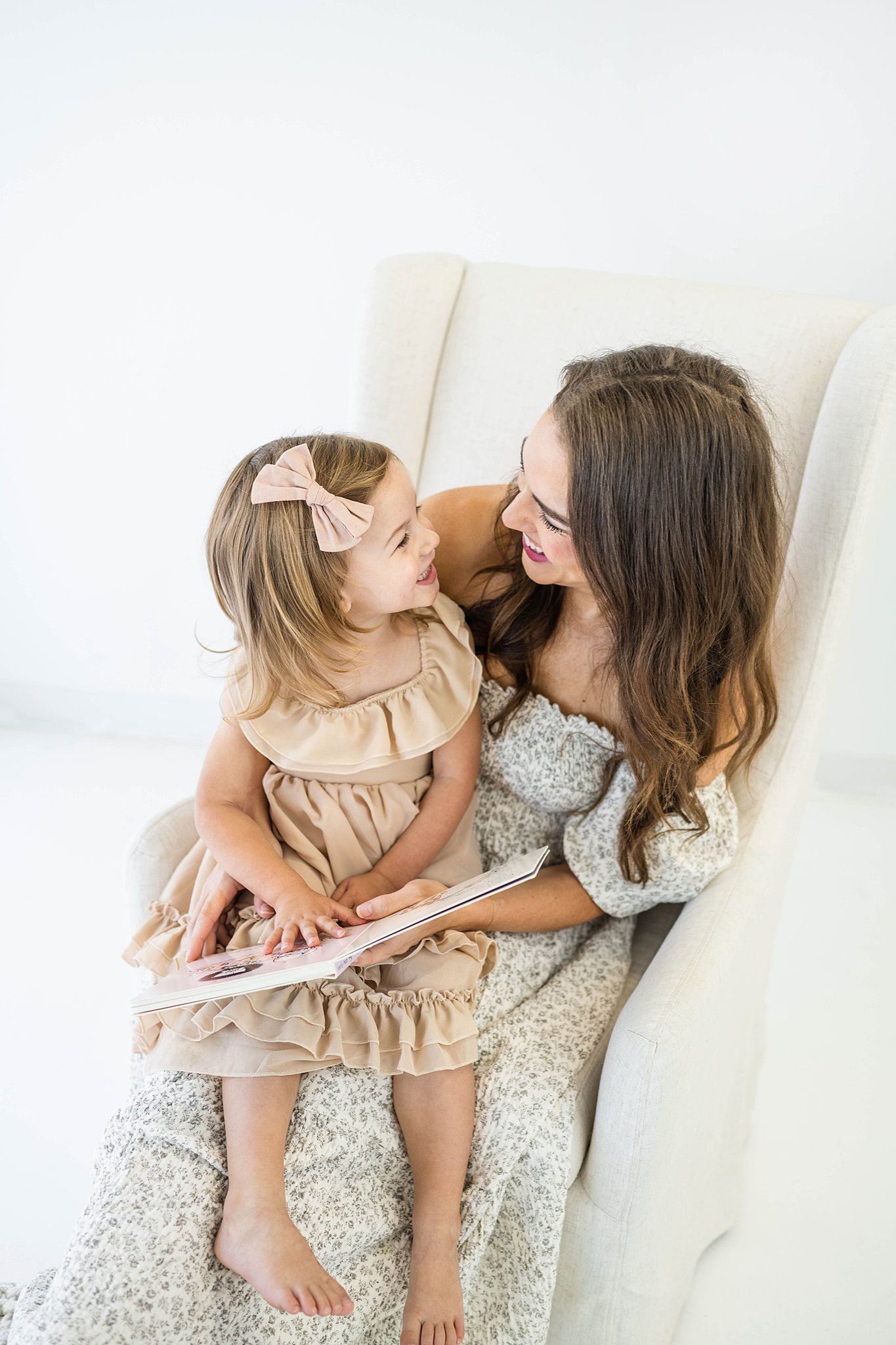 A mother in a pattern dress sits in a white chair with her toddler daughter wearing a beige dress reading a book together mommy and me classes okc