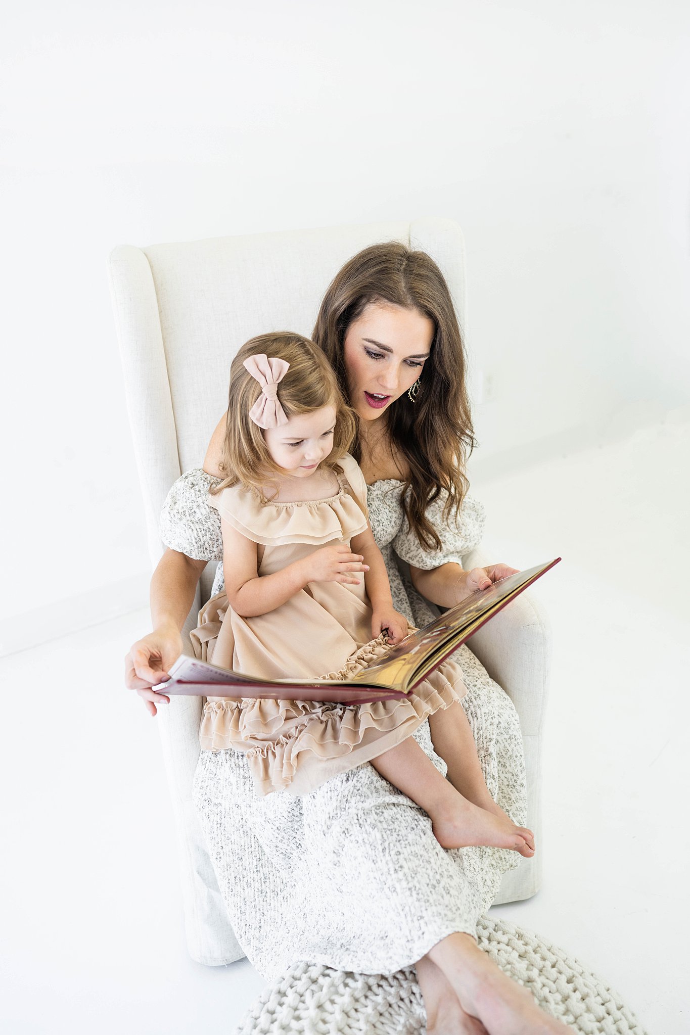 A mother and young daughter sit in a white chair reading a book together in a studio mommy and me classes okc