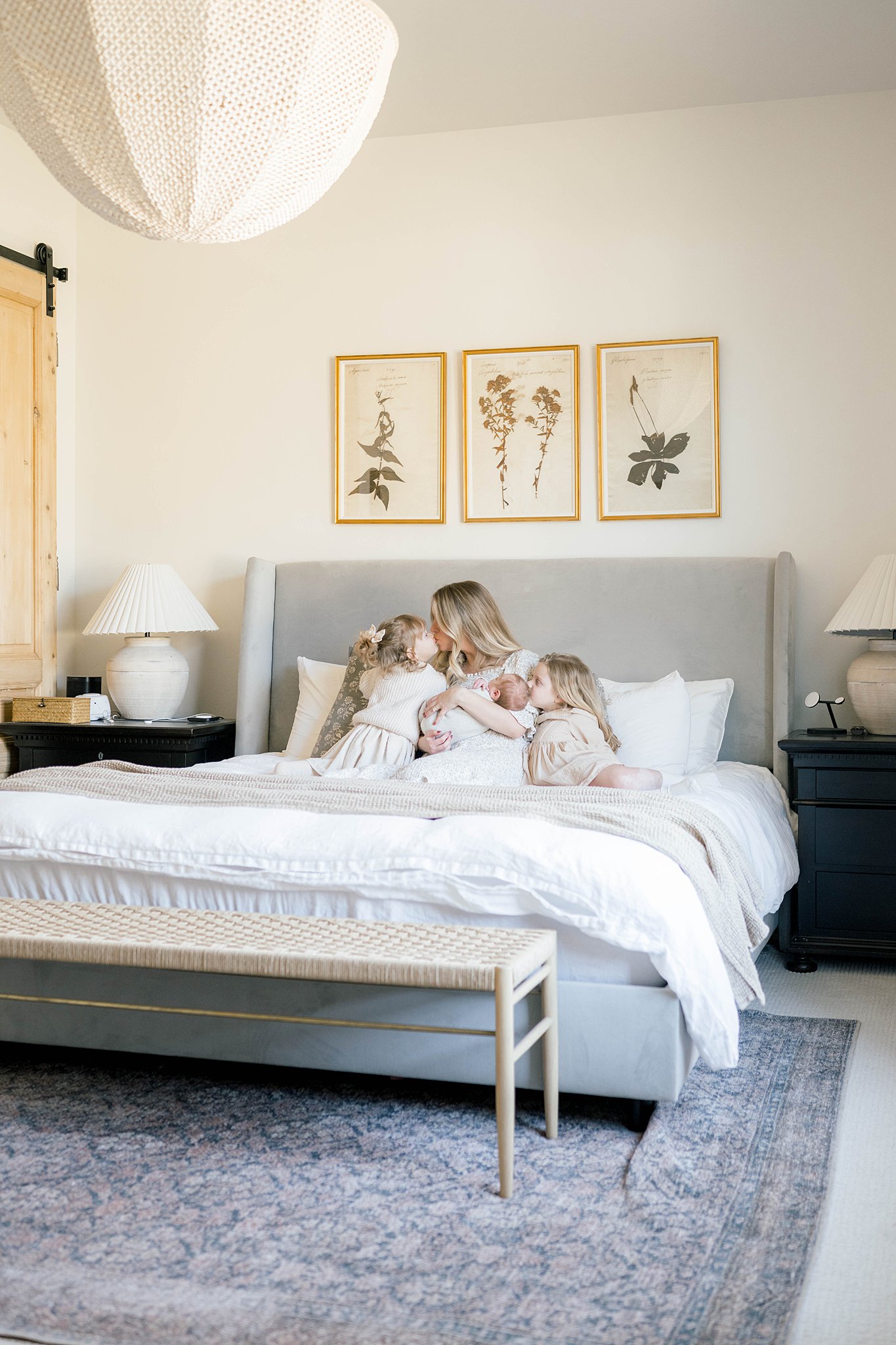 A mother sits on a bed holding her newborn with two daughters kissing one of her daughters with a grey bed and floral paintings above them okc interior design