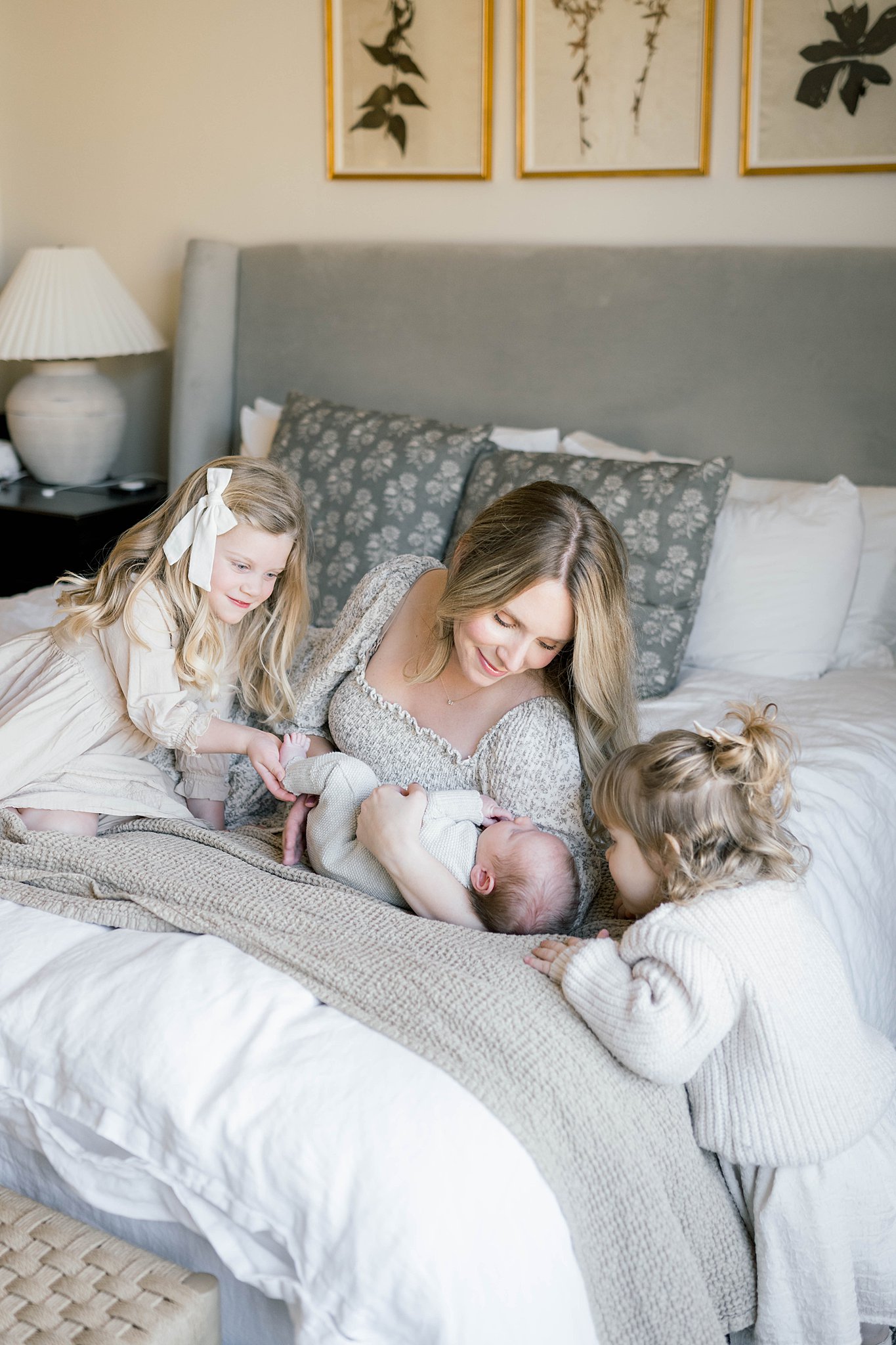 A mother lays across a grey and white bed cuddling her newborn baby while her other two daughters look on from either side okc interior design