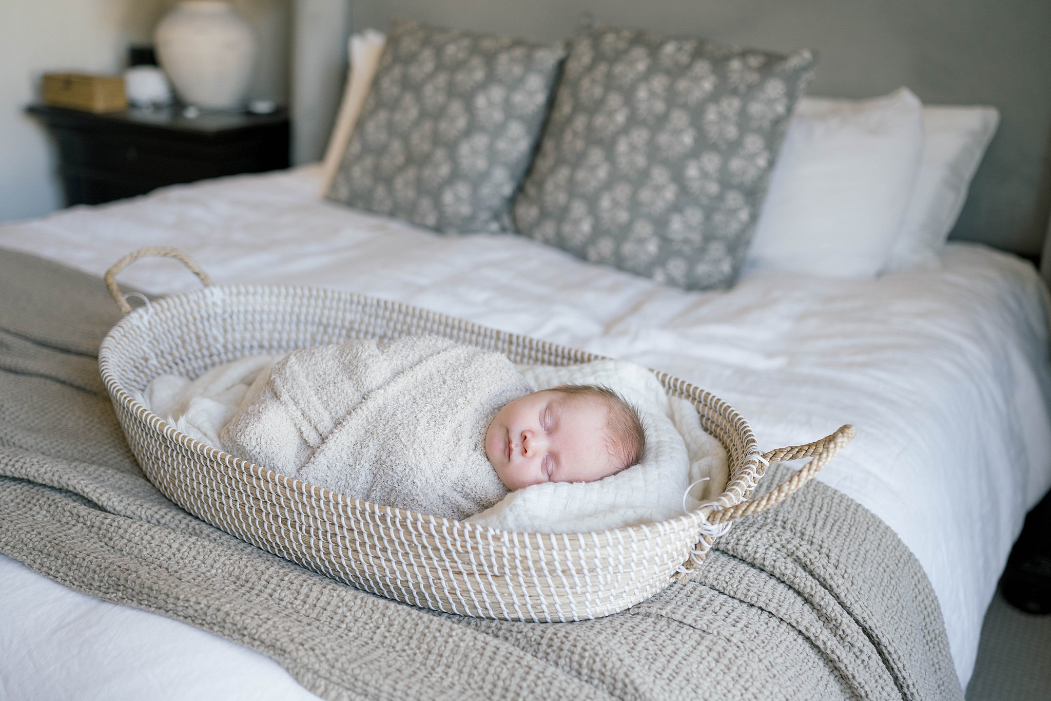 A newborn baby swaddled in a beige blanket sleeps in a long basket on a bed
