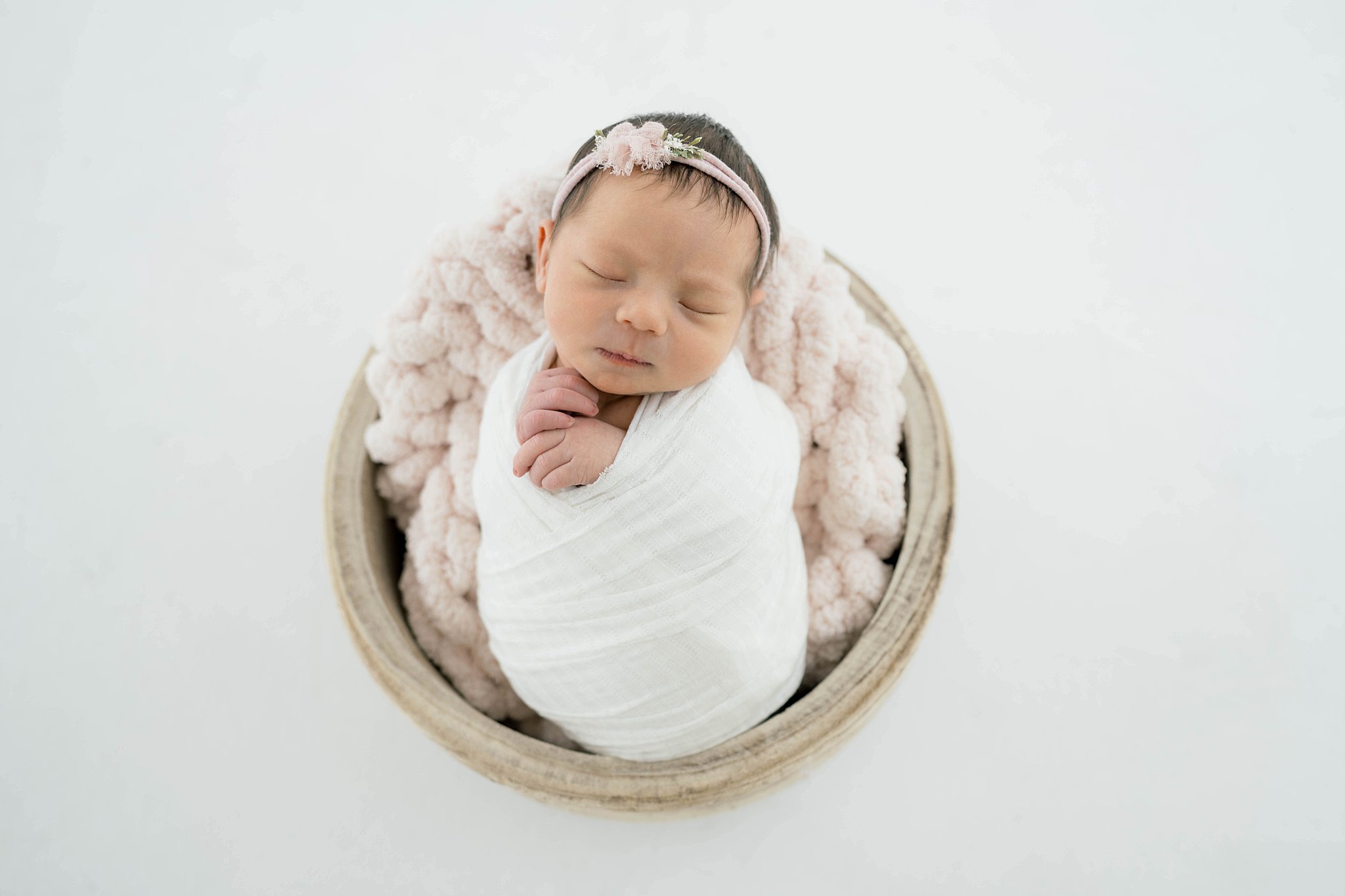 A newborn baby in a white swaddle sleeps on a pink blanket in a wooden bucket