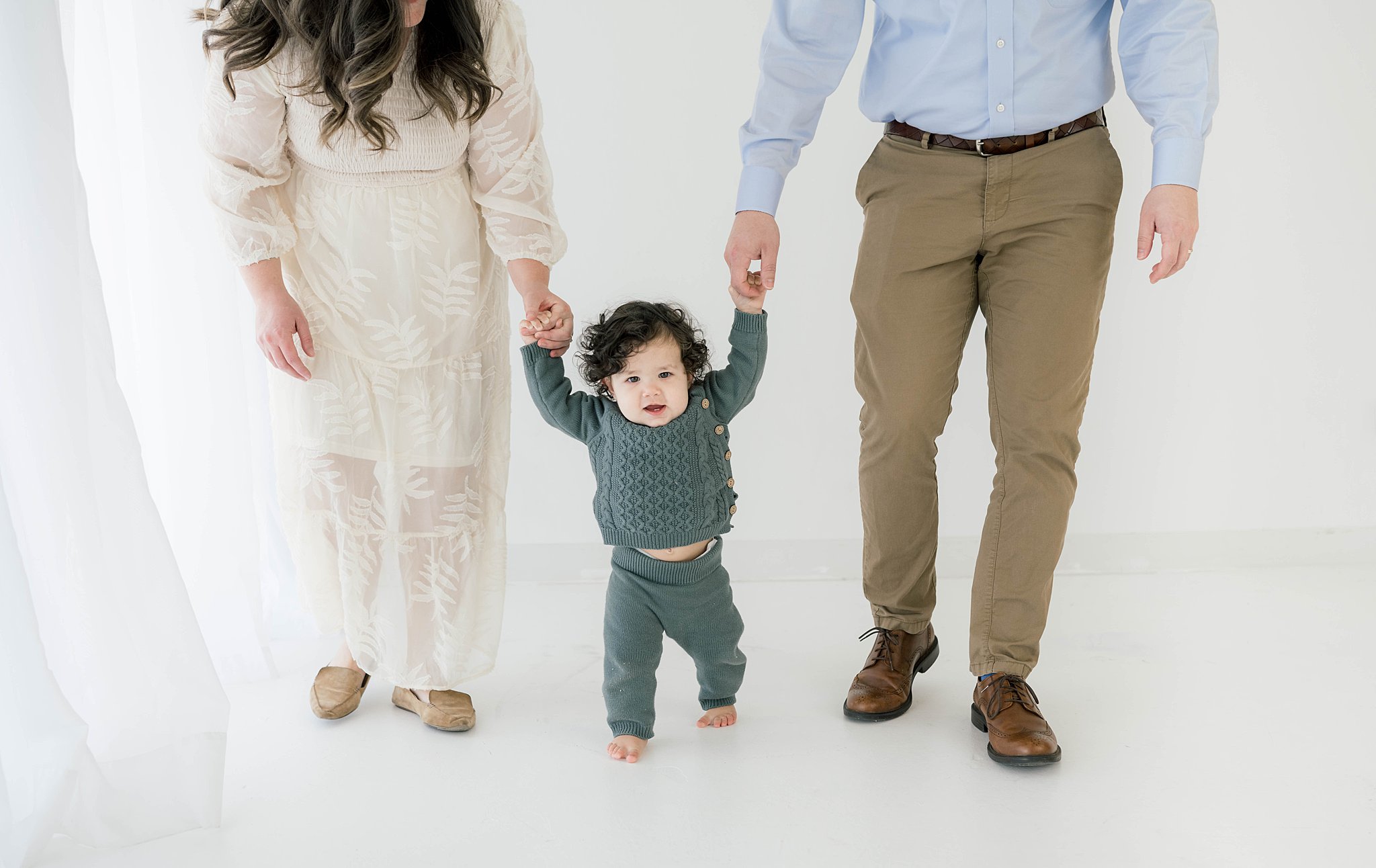 A mother in a white lace dress and father in khaki pants hold the hands of their toddler son while walking through a studio oklahoma city pediatricians