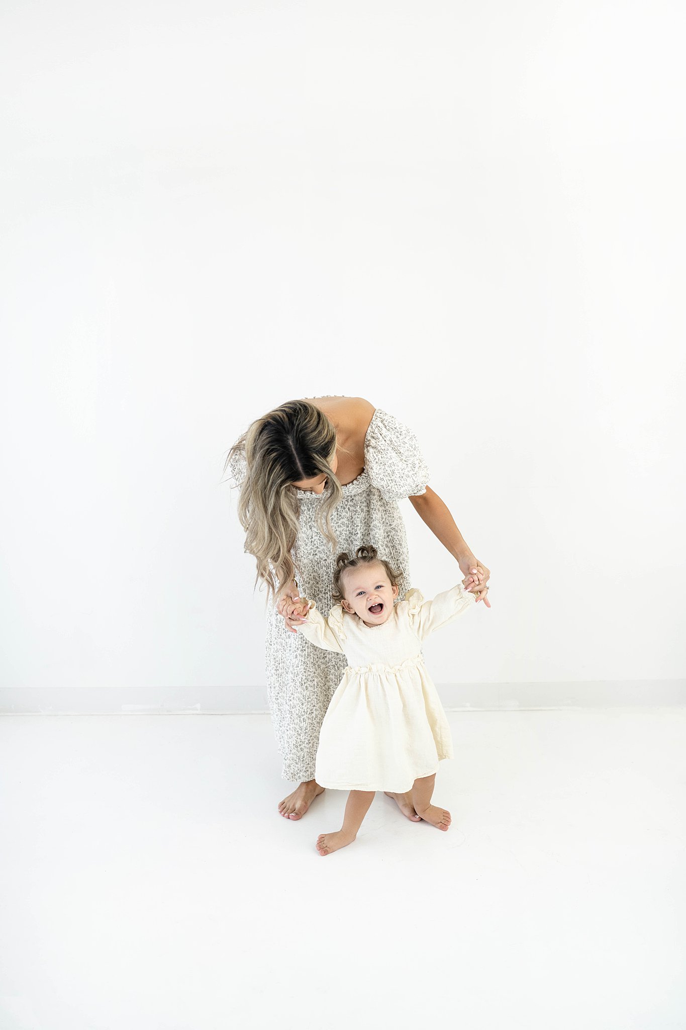 A mother stands in a studio holding the hands and playing with her toddler daughter