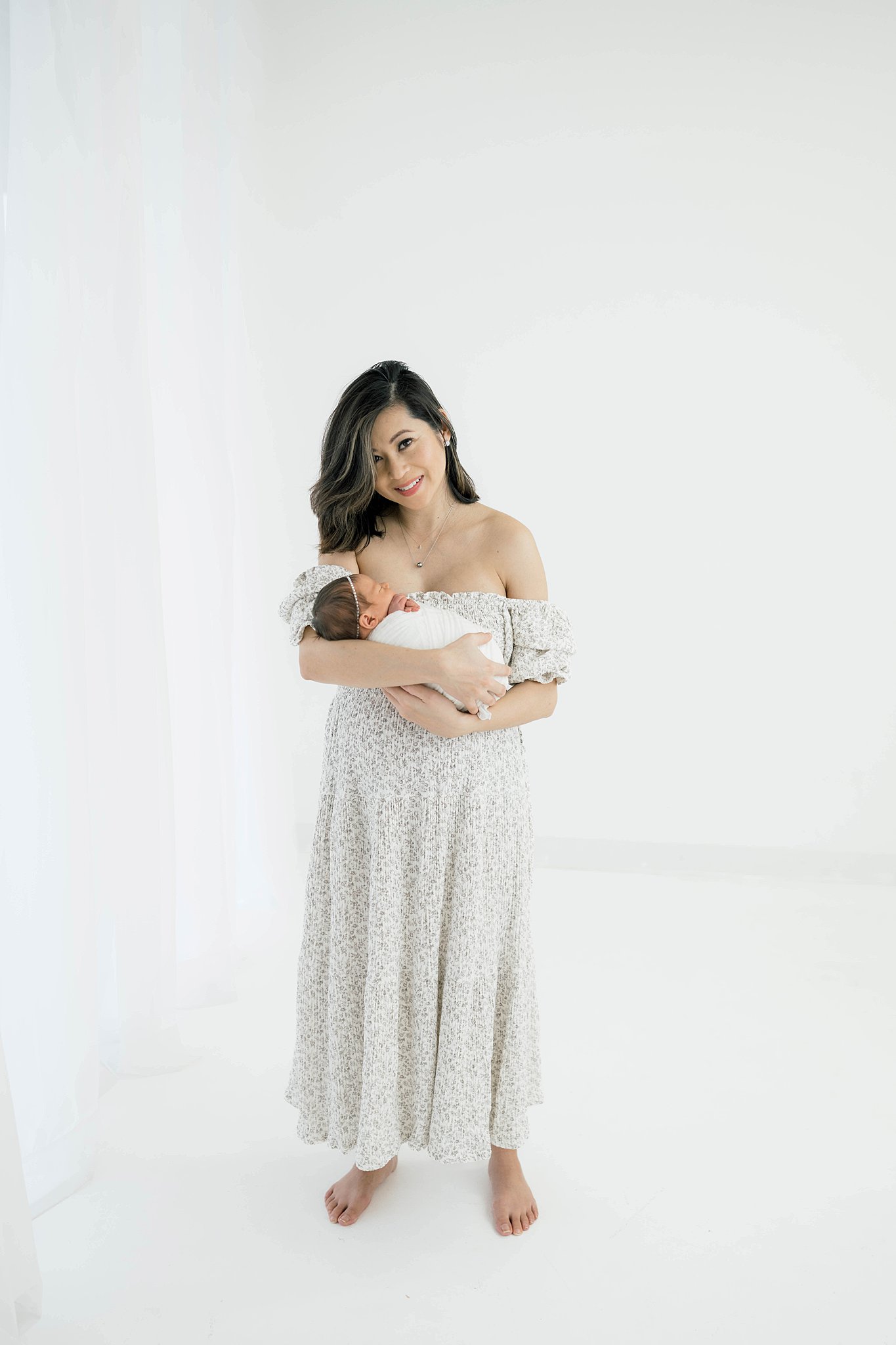 A mother in an off the shoulder dress stands in a studio by a window holding her newborn baby daughter pelvic floor therapy okc