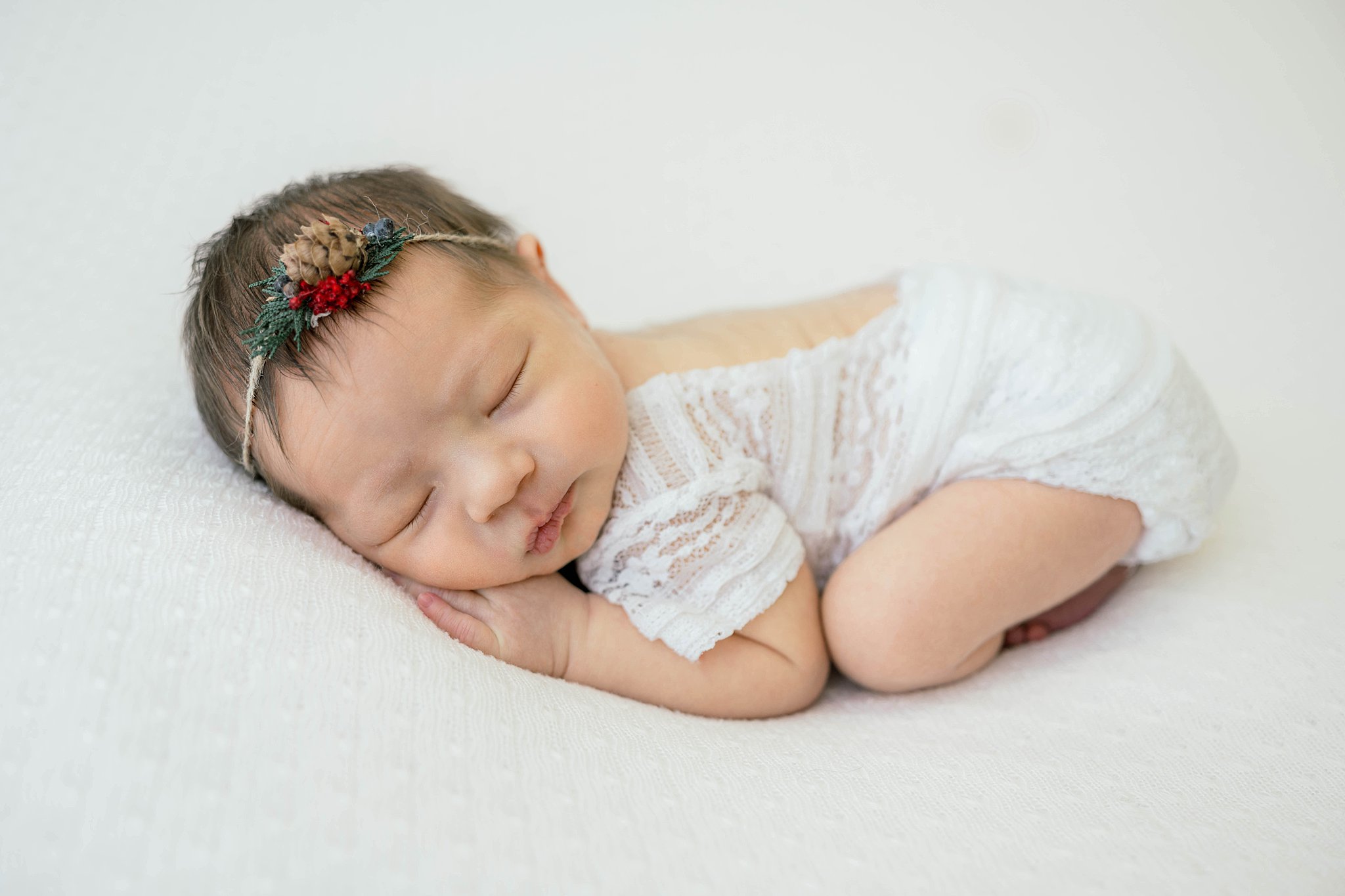 A newborn baby sleeps in a lace onesie while wearing a christmas wreath headband ultrasound oklahoma city