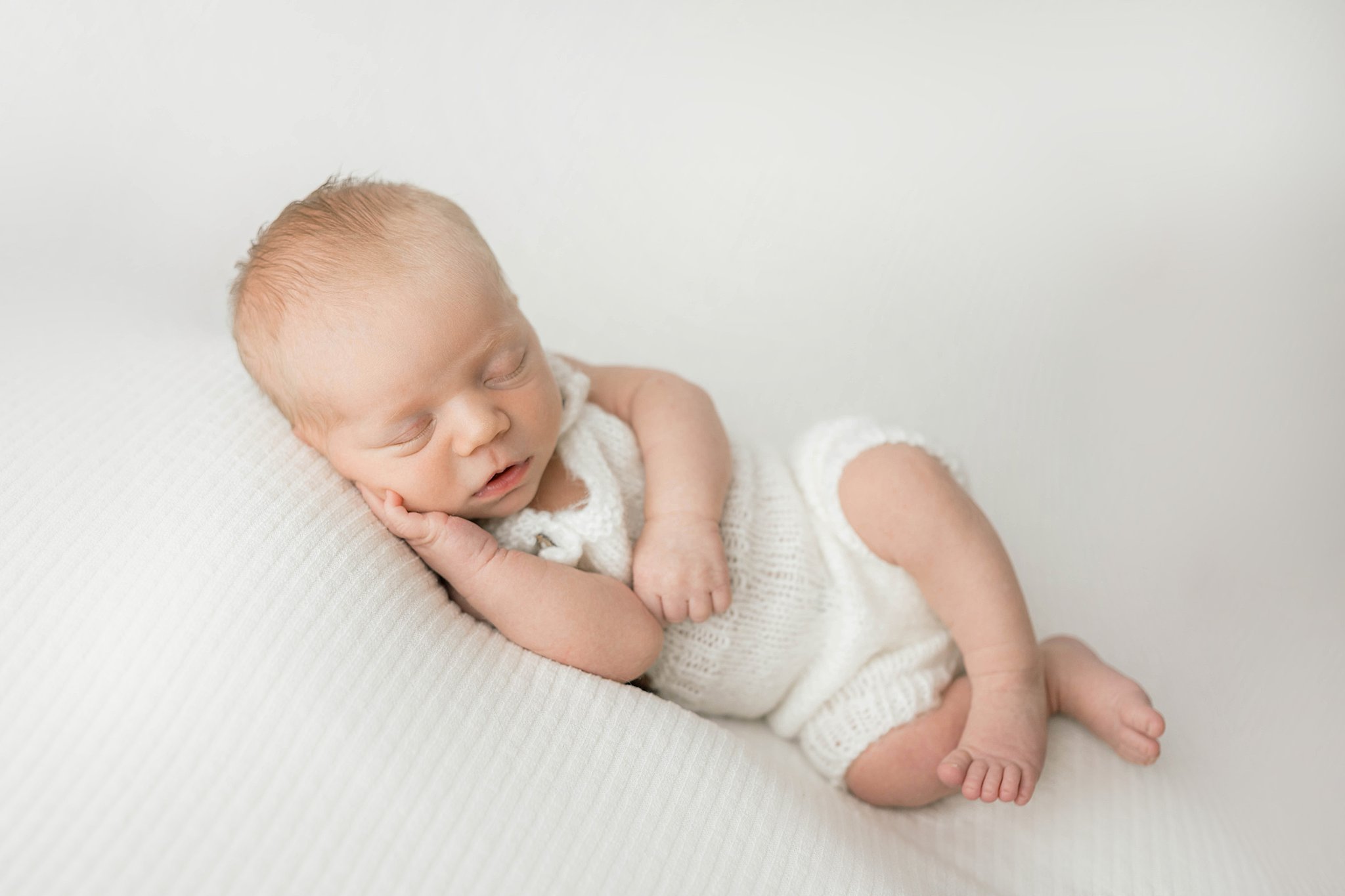 A newborn baby lounges and sleeps in a white knit onesie
