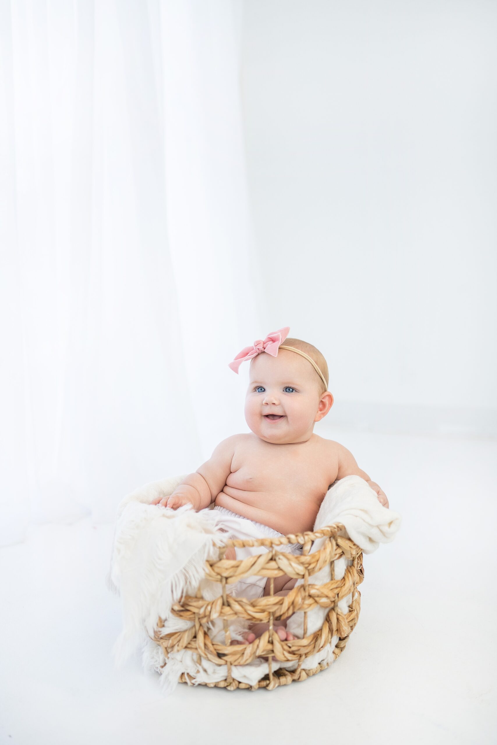 An infant baby with a pink bow sits in a woven basket with a white blanket in a studio natural okie baby