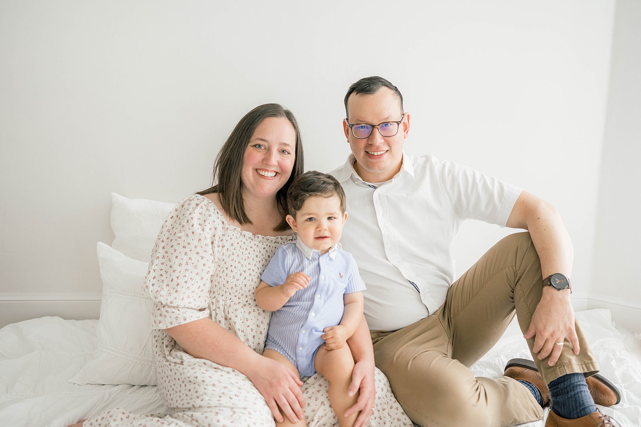 Mom and dad sit on a white bed while their toddler son in a blue onesie sits on mom's lap