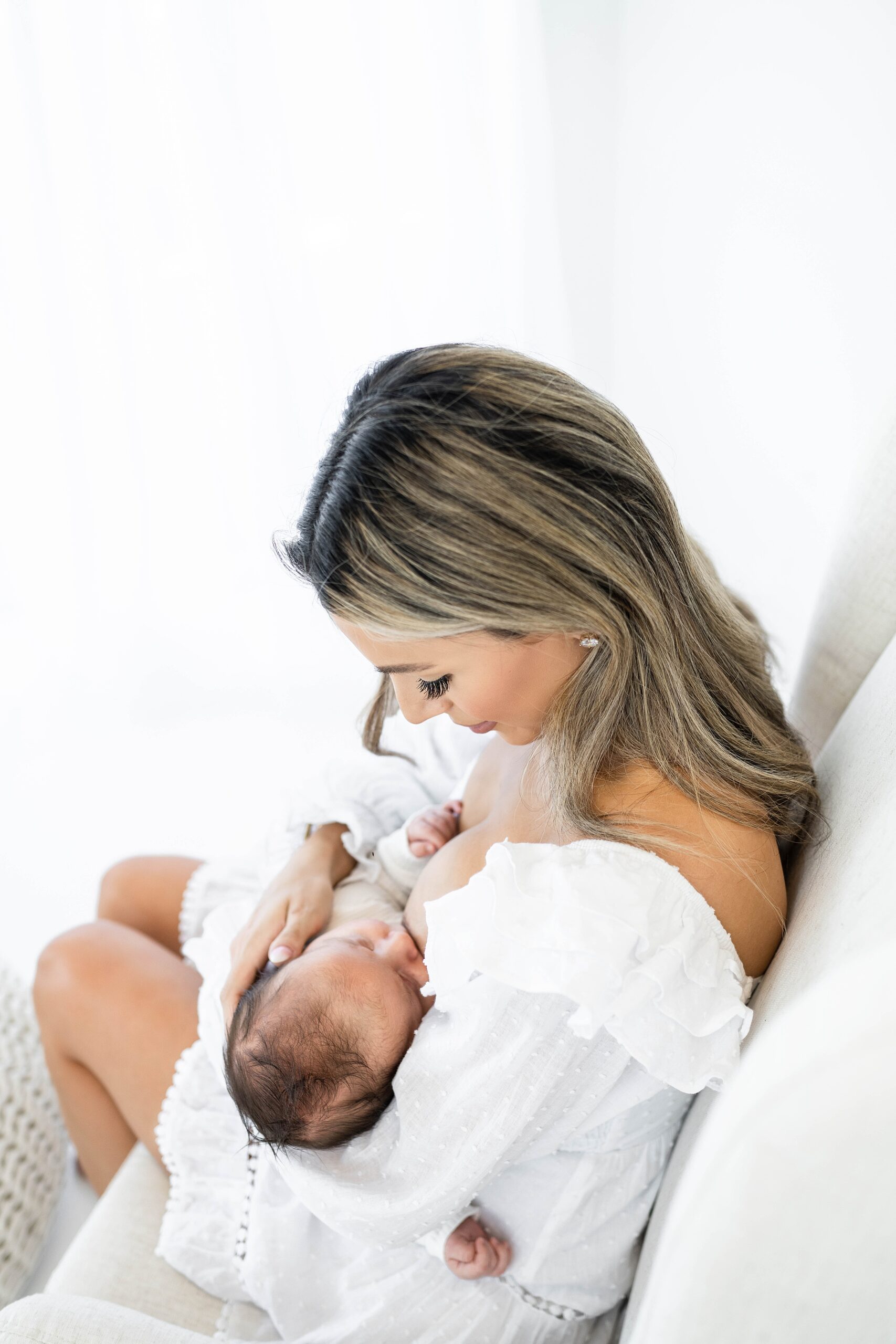 A mother to be breast feeds her newborn baby while sitting in a white chair in a studio