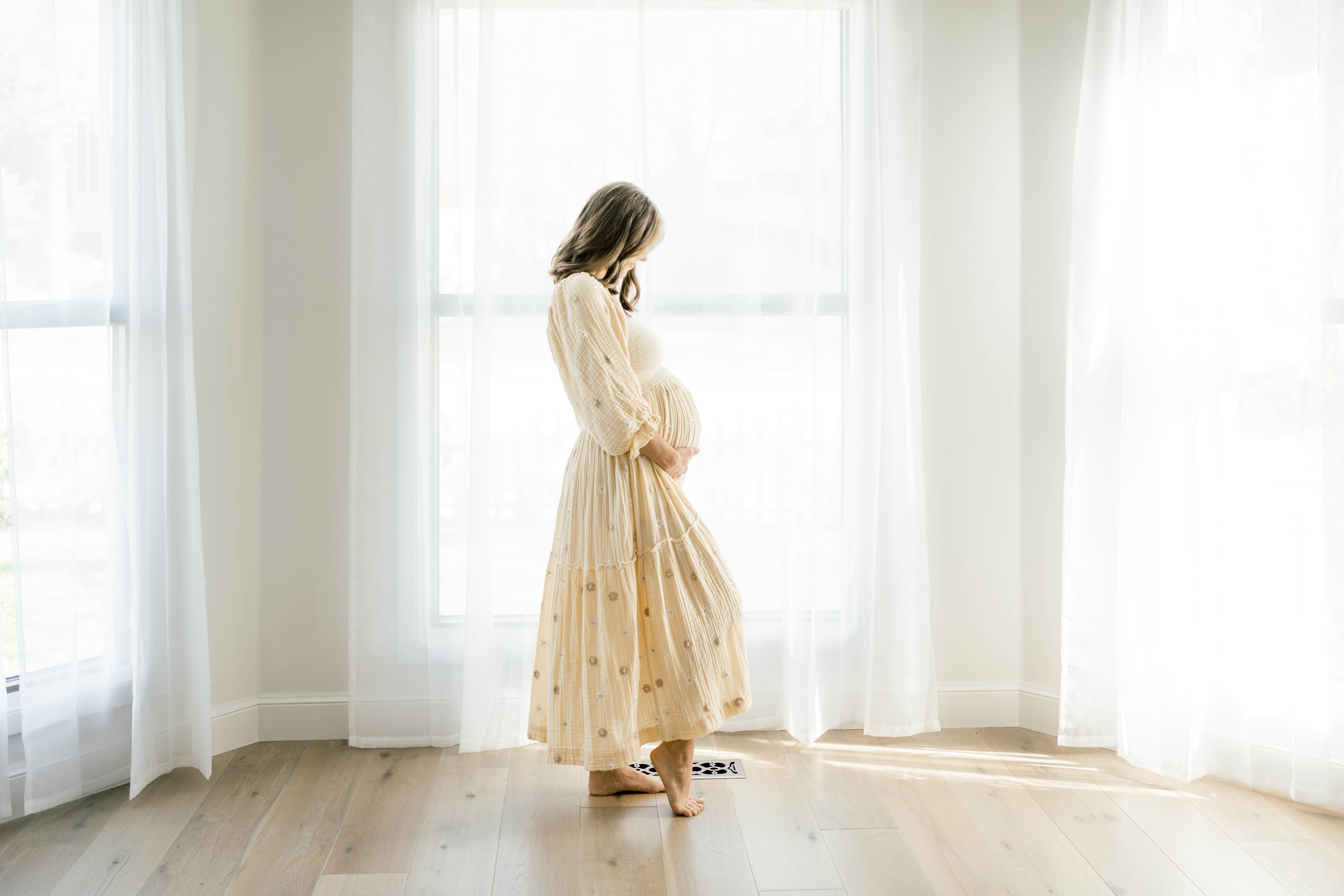 A mom to be in a yellow maternity dress stands in front of windows looking down at the bump