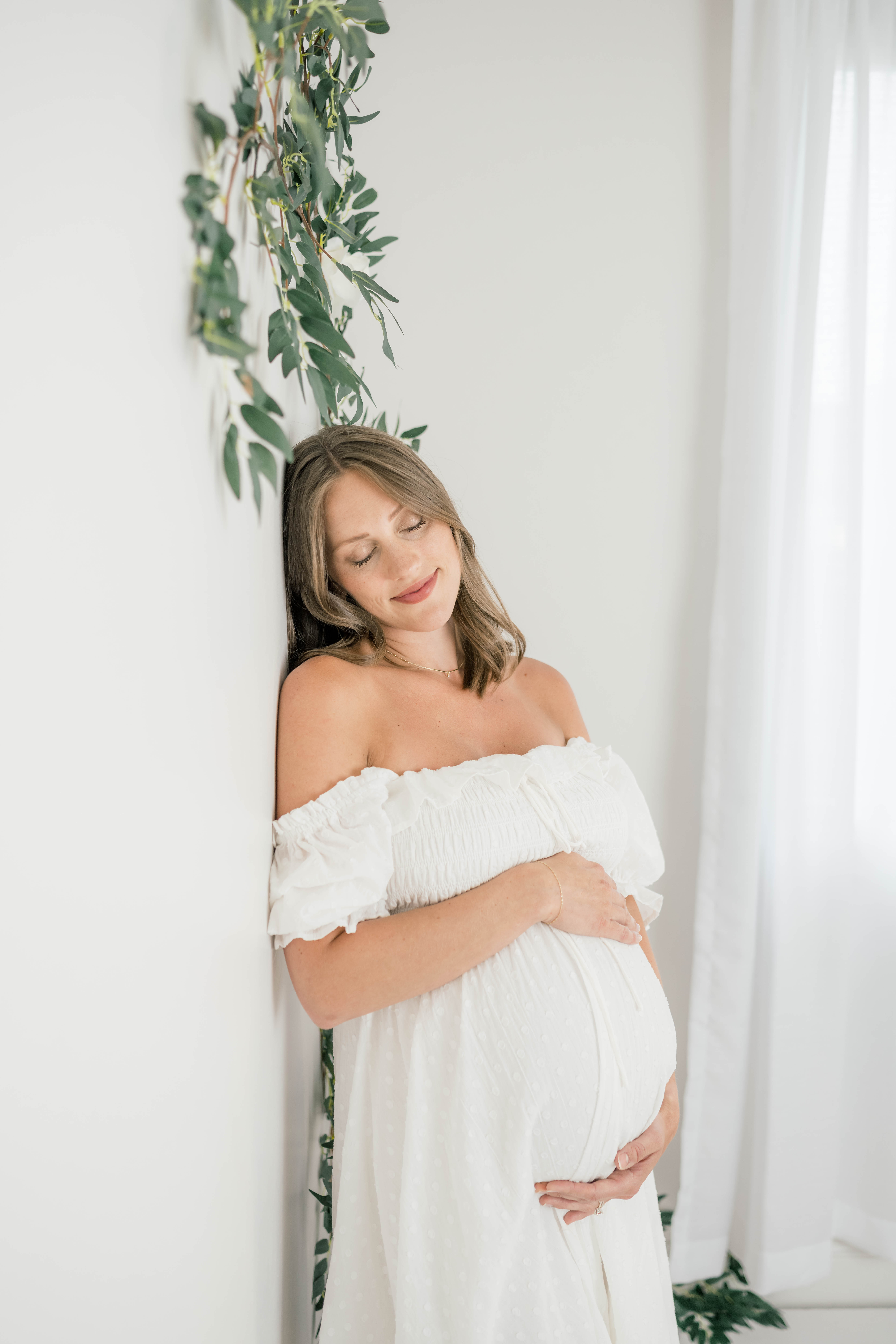 A mom to be leans against a wall covered in greenery while holding her bump in a white maternity gown the nest okc
