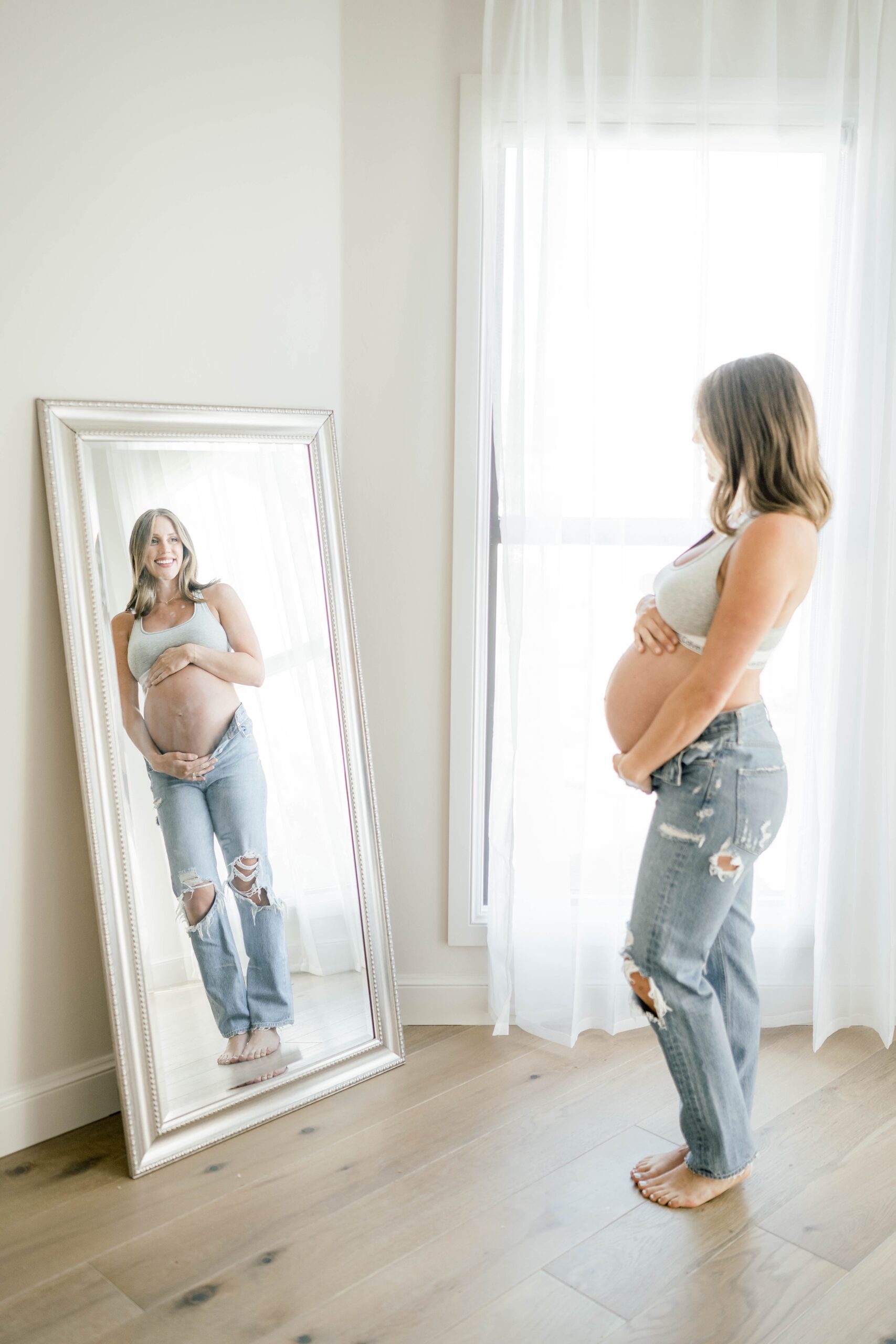 A mother to be stands in a studio in front of a mirror wearing jeans and sports bra water birth okc