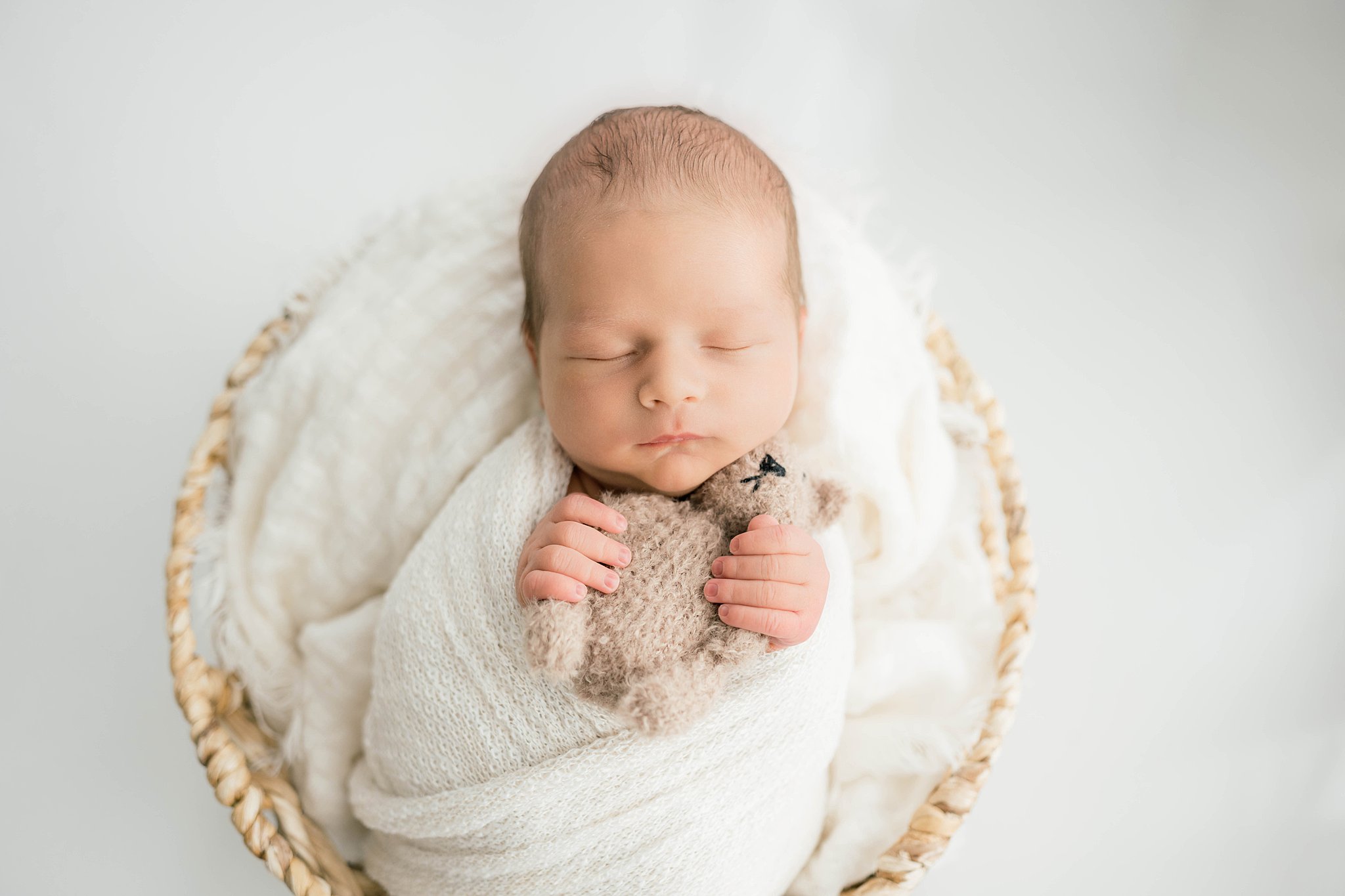 A newborn baby sleeps in a white swaddle in a woven basket cradling a small crochet stuffed animal kidzone furniture