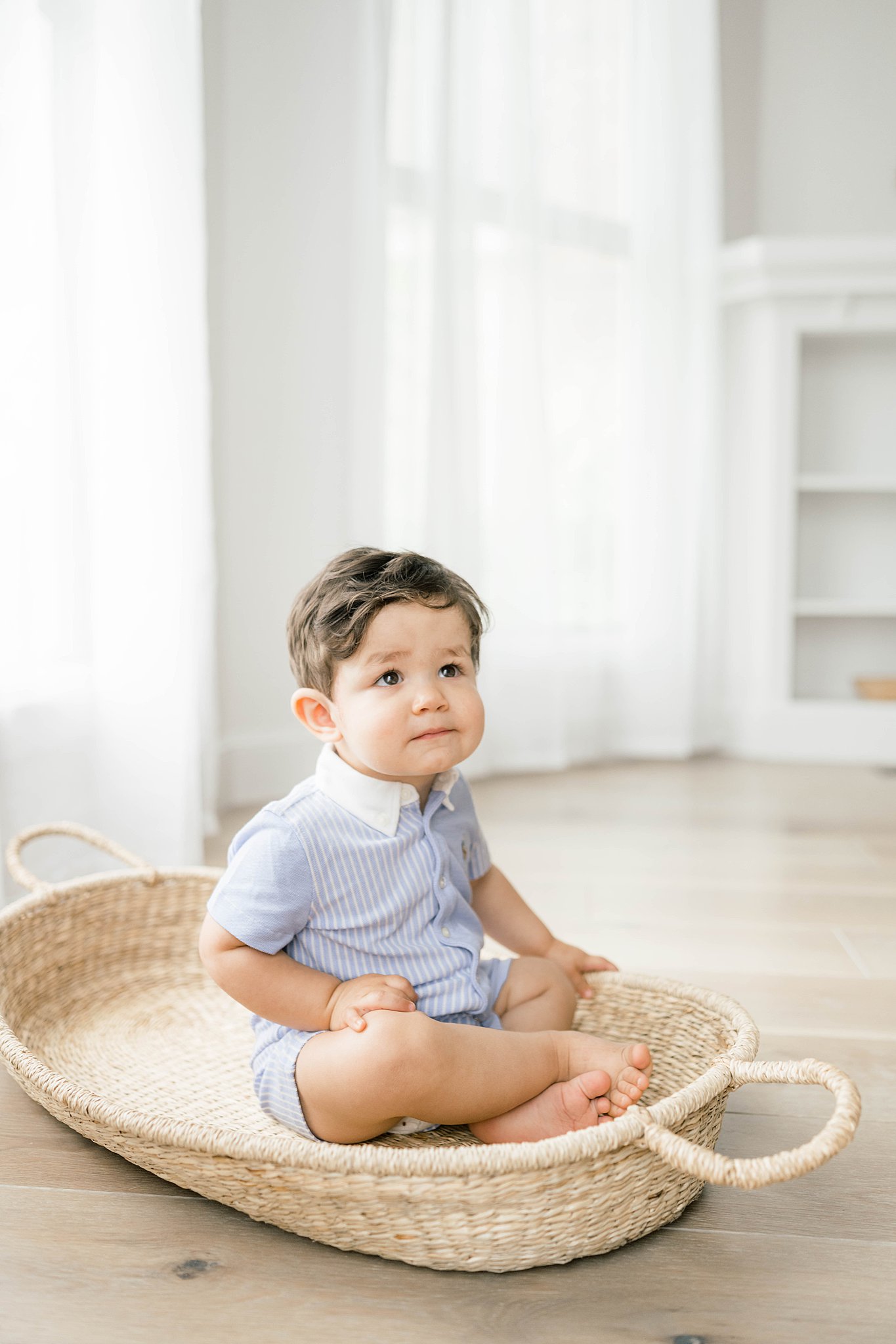A young boy sits in a woven Moses basket in a blue onesie with stripes and a white collar in a studio okc indoor playground