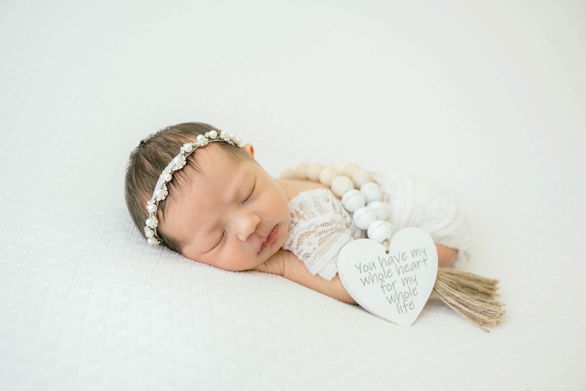 A newborn baby sleeps in a lace onesie with a heart necklace draped over her