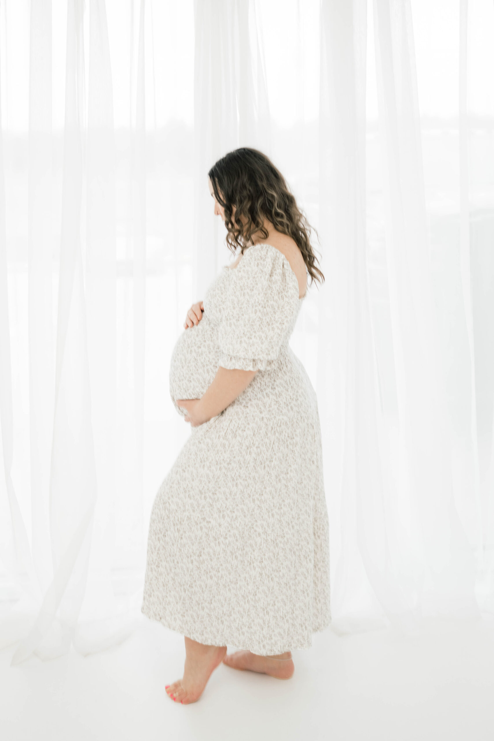 A mother to be stands by a window looking down at her bump with her hands placed on it prenatal chiropractor okc