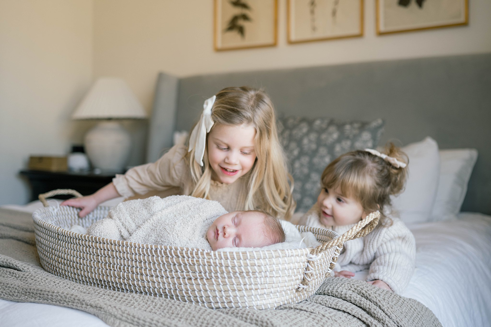 Two young sisters look over their sleeping newborn baby sibling in a woven basket on a bed