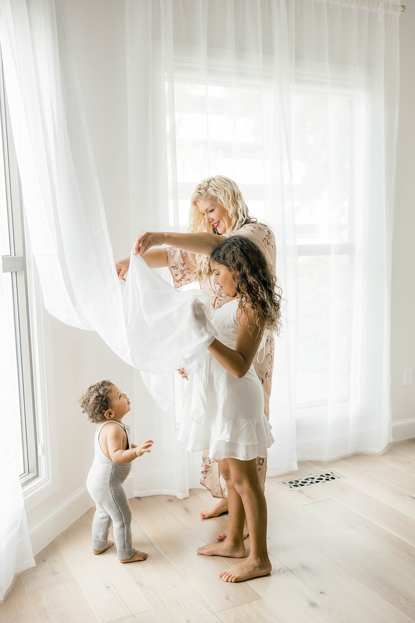 A mother plays in a window curtain with her toddler and young daughter