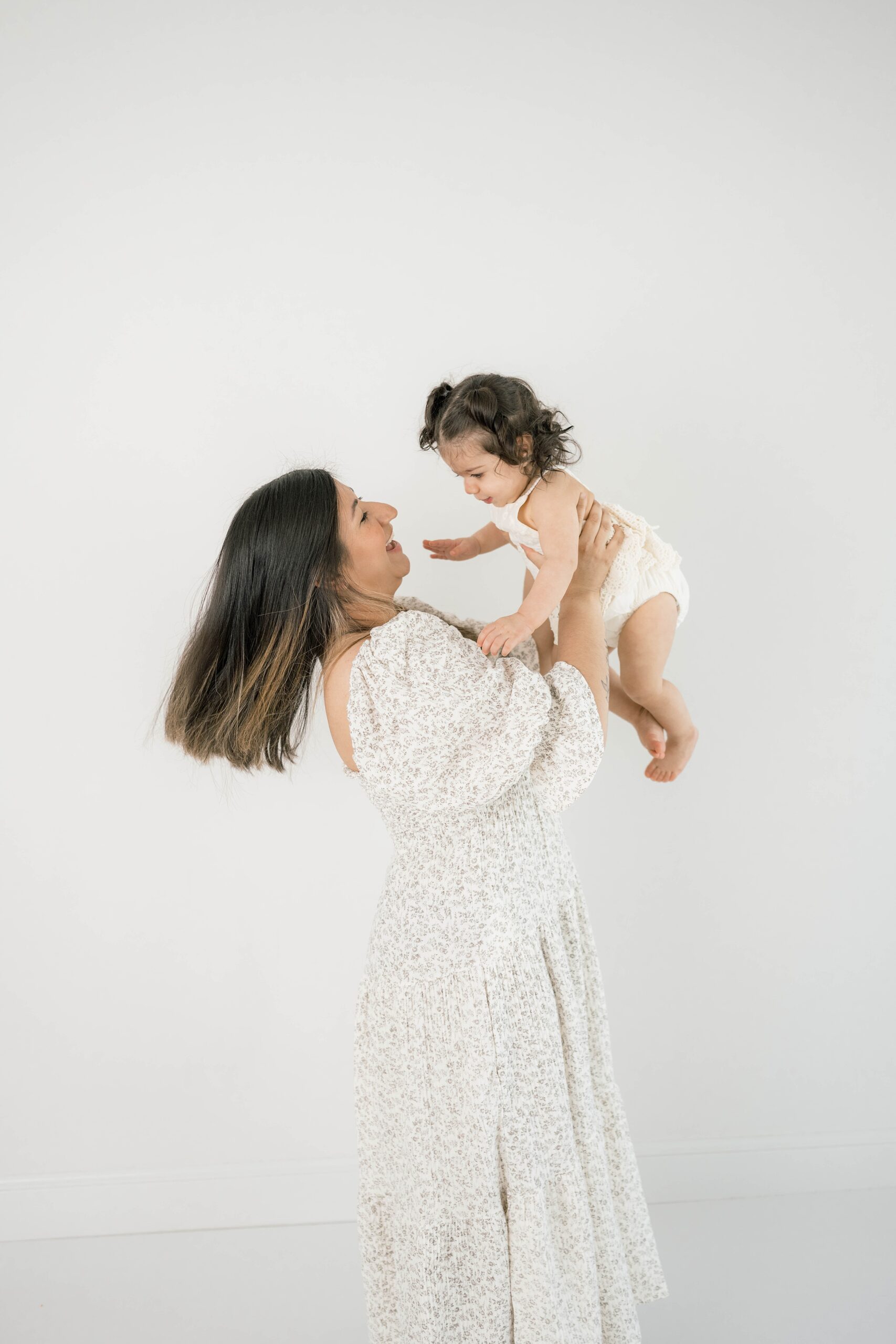 A mother in a white floral print dress lifts and plays with her toddler daughter in a studio