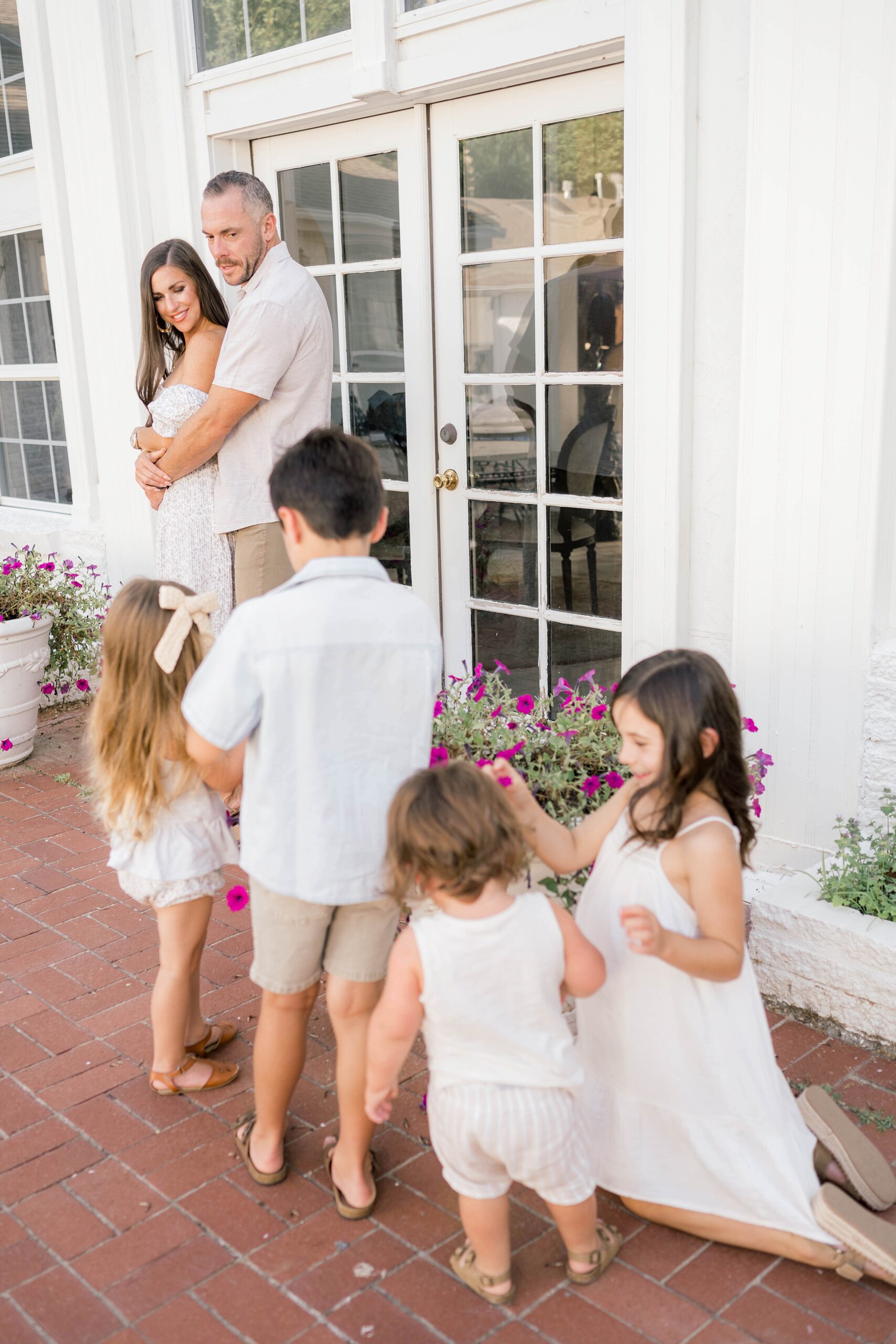 A mom and dad hug while their four young children play with flowers on a brick patio in white clothes before visiting kid friendly restaurants okc