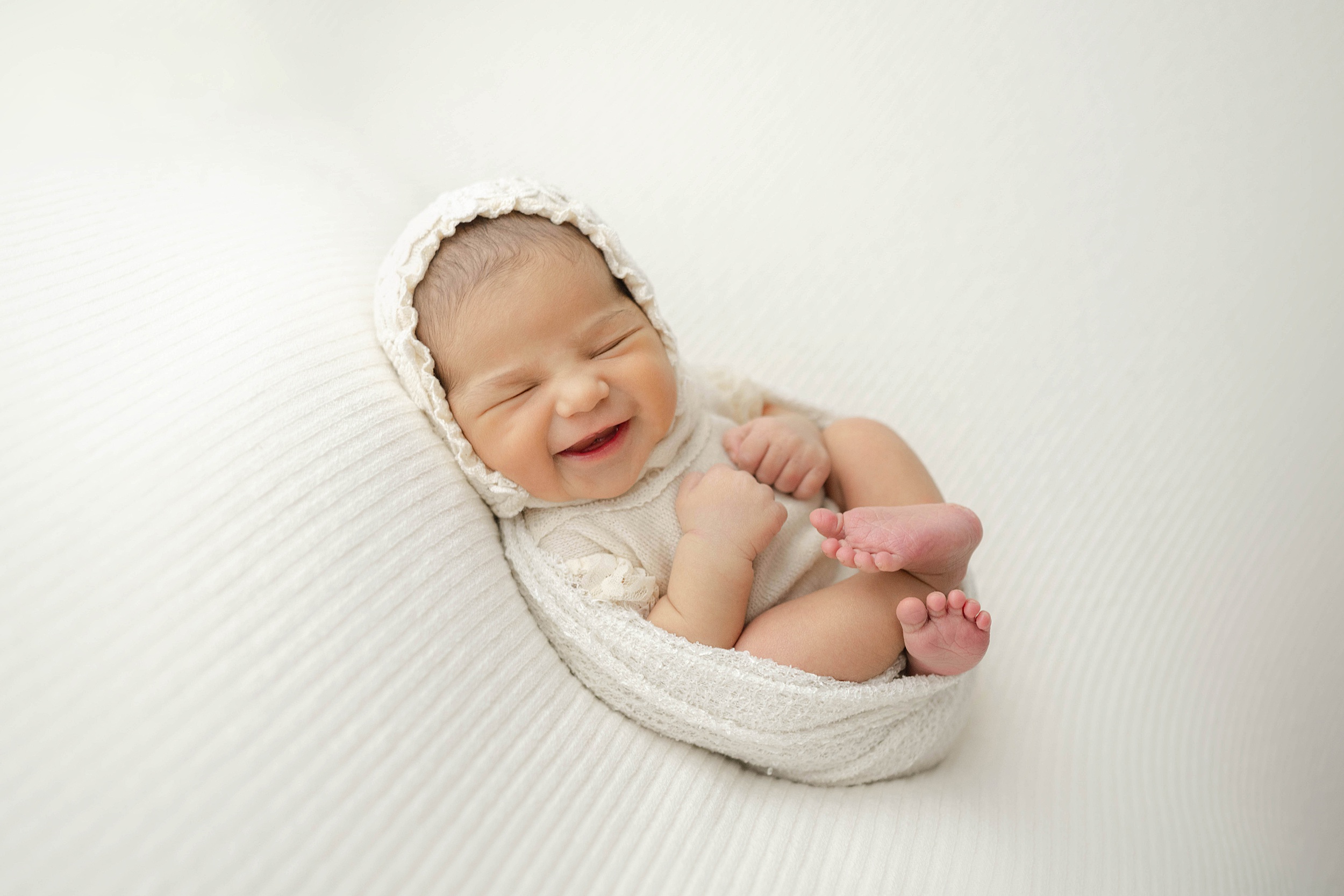 A newborn baby smiles in its sleep while wrapped in a cheesecloth swaddle and white bonnet