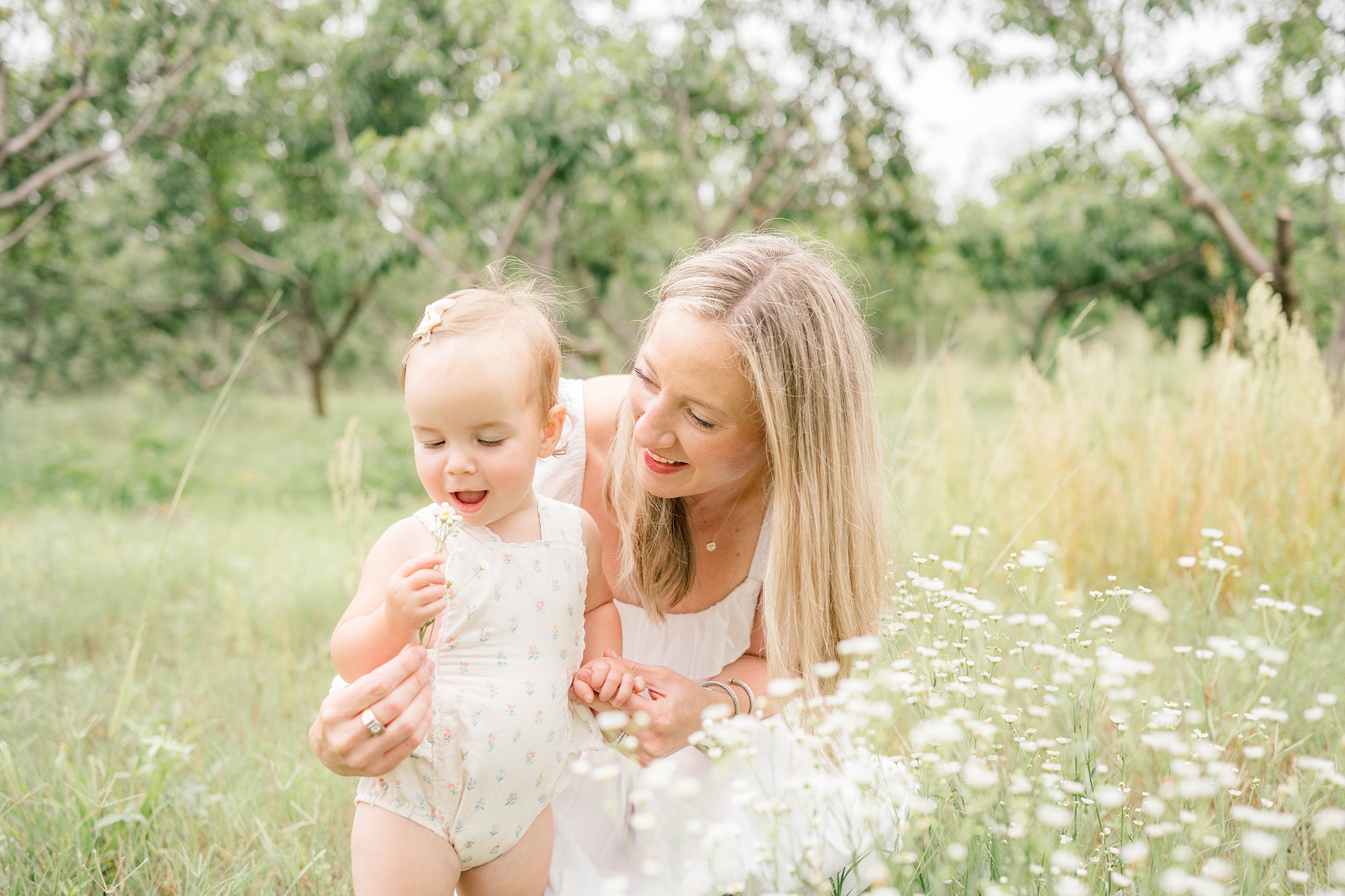 A mother in a white dress plays with her toddler daughter exploring white wildflowers after visiting oklahoma city preschools