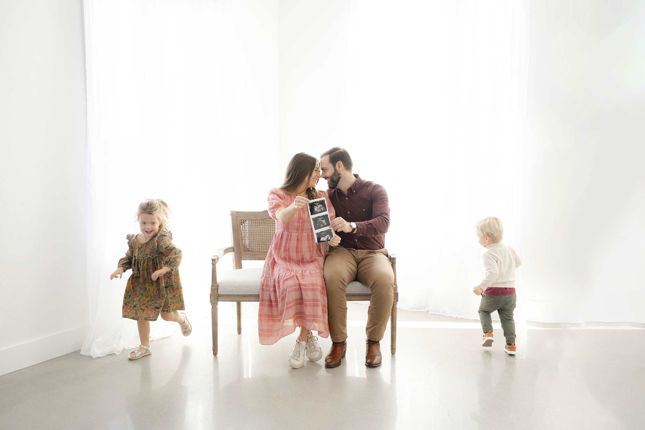 Pregnant parents sit on a bench holding their sonogram while their toddler son and daughter run circles around them in a studio