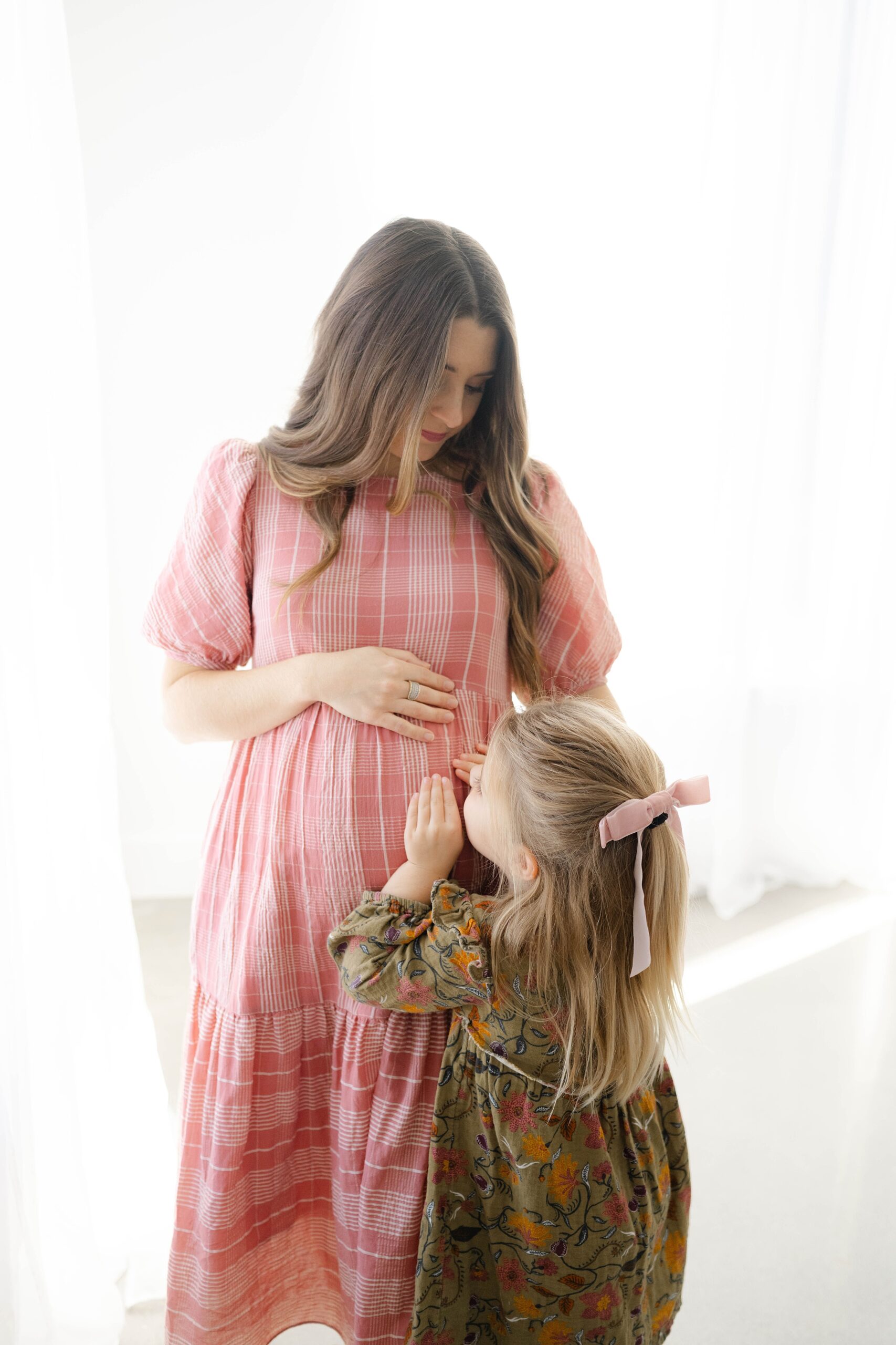 A pregnant mother in a pink plaid dress stands in a window of a studio while her toddler daughter kisses the bump after using ou fertility clinic OKC