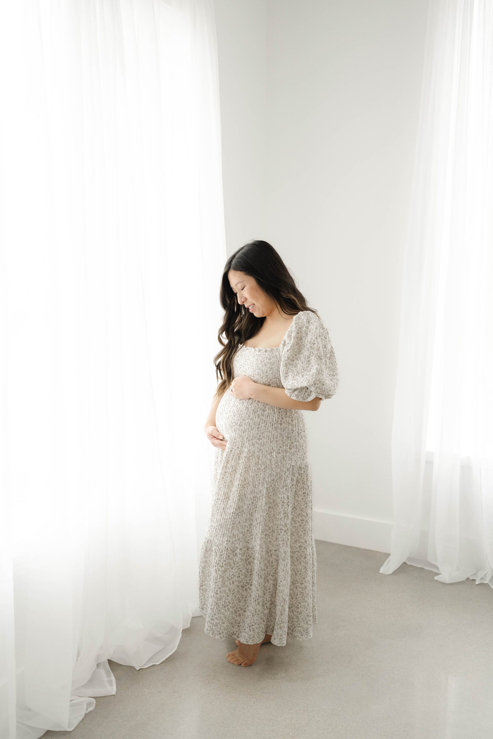 A mother in a floral dress stands in a window of a studio smiling down at the bump in her hands thanks to ou obgyn
