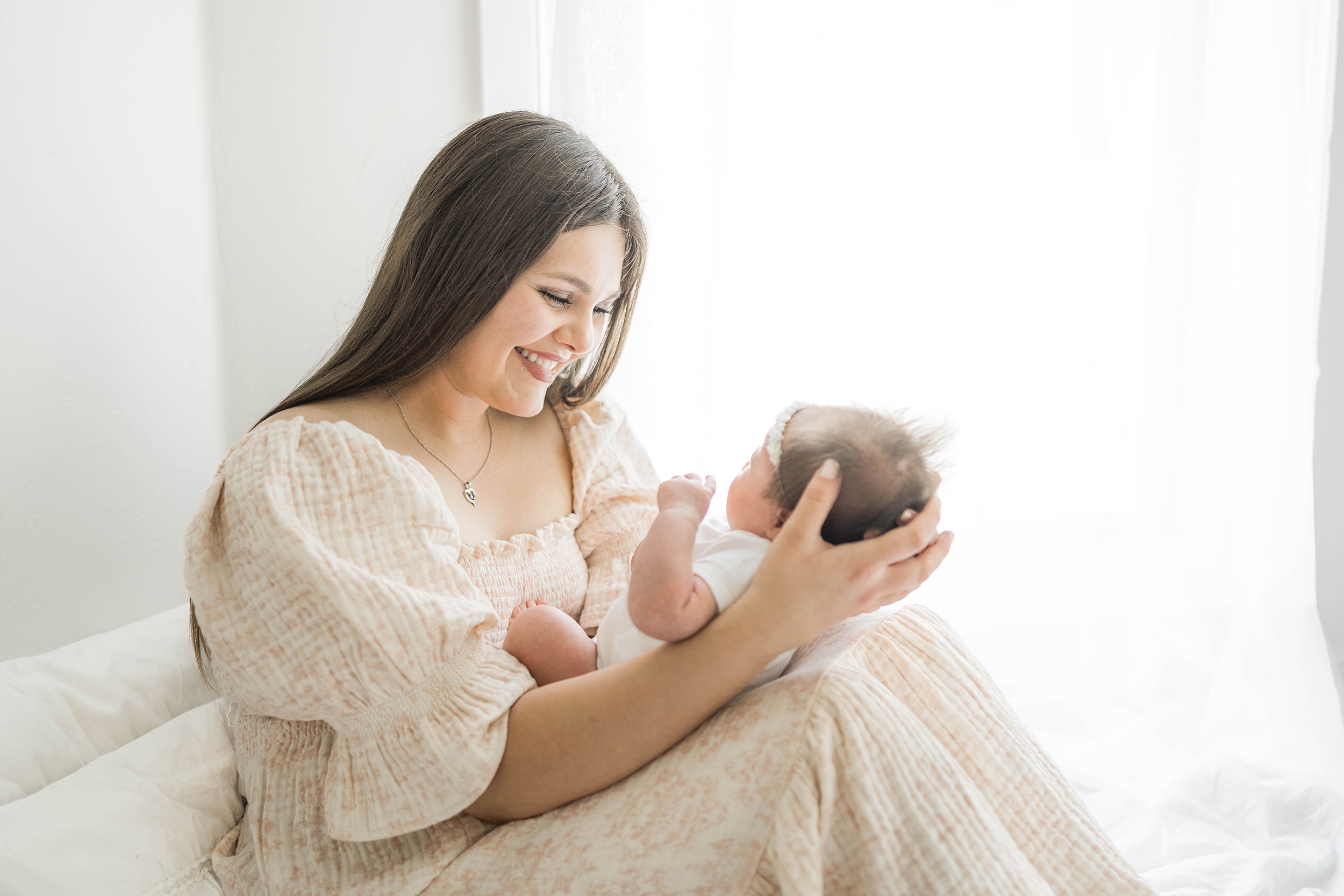 A mother in a floral dress sits on a bed smiling down at her newborn baby daughter in her lap