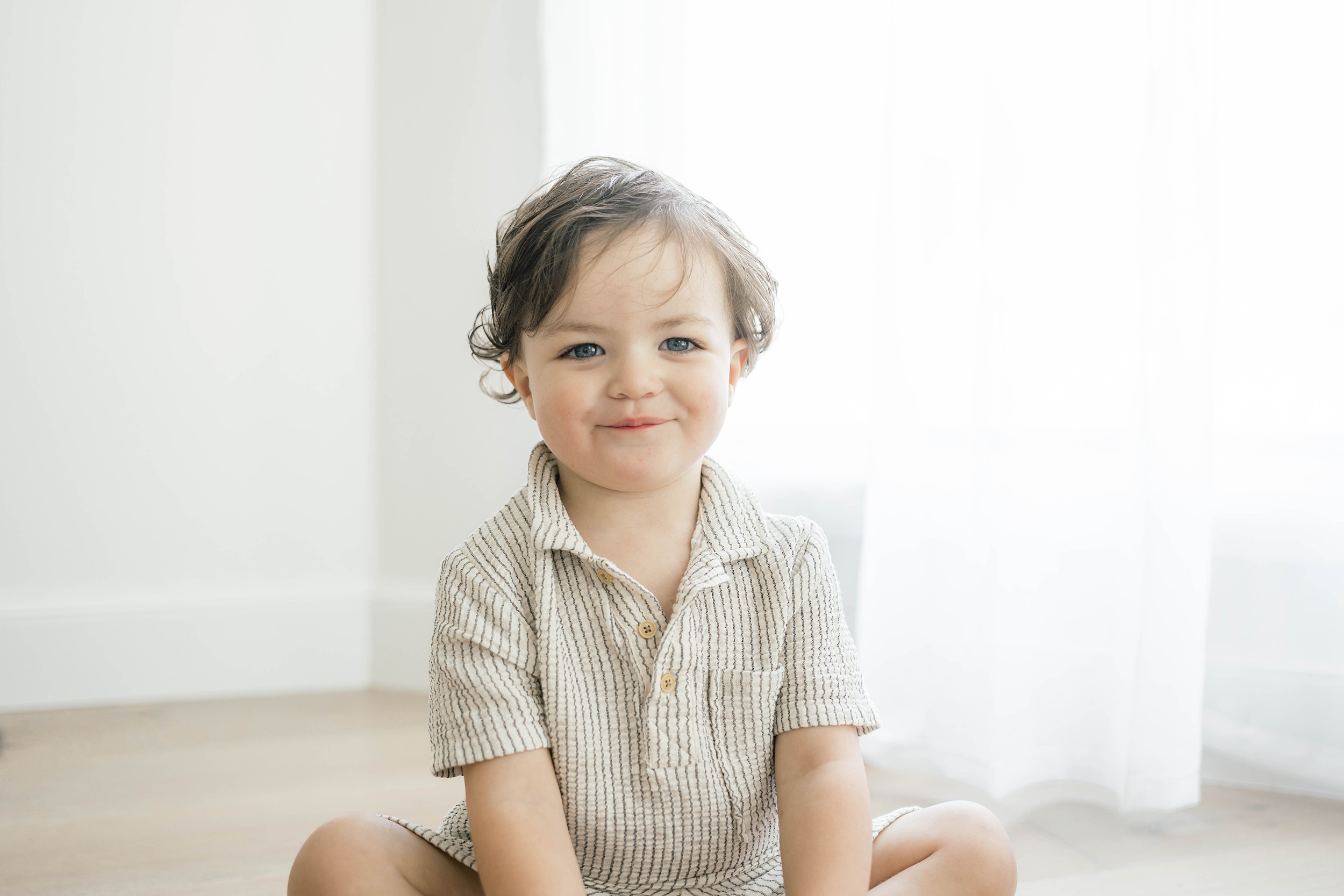 A young toddler in a striped shirt sits on a studio floor by a window smiling