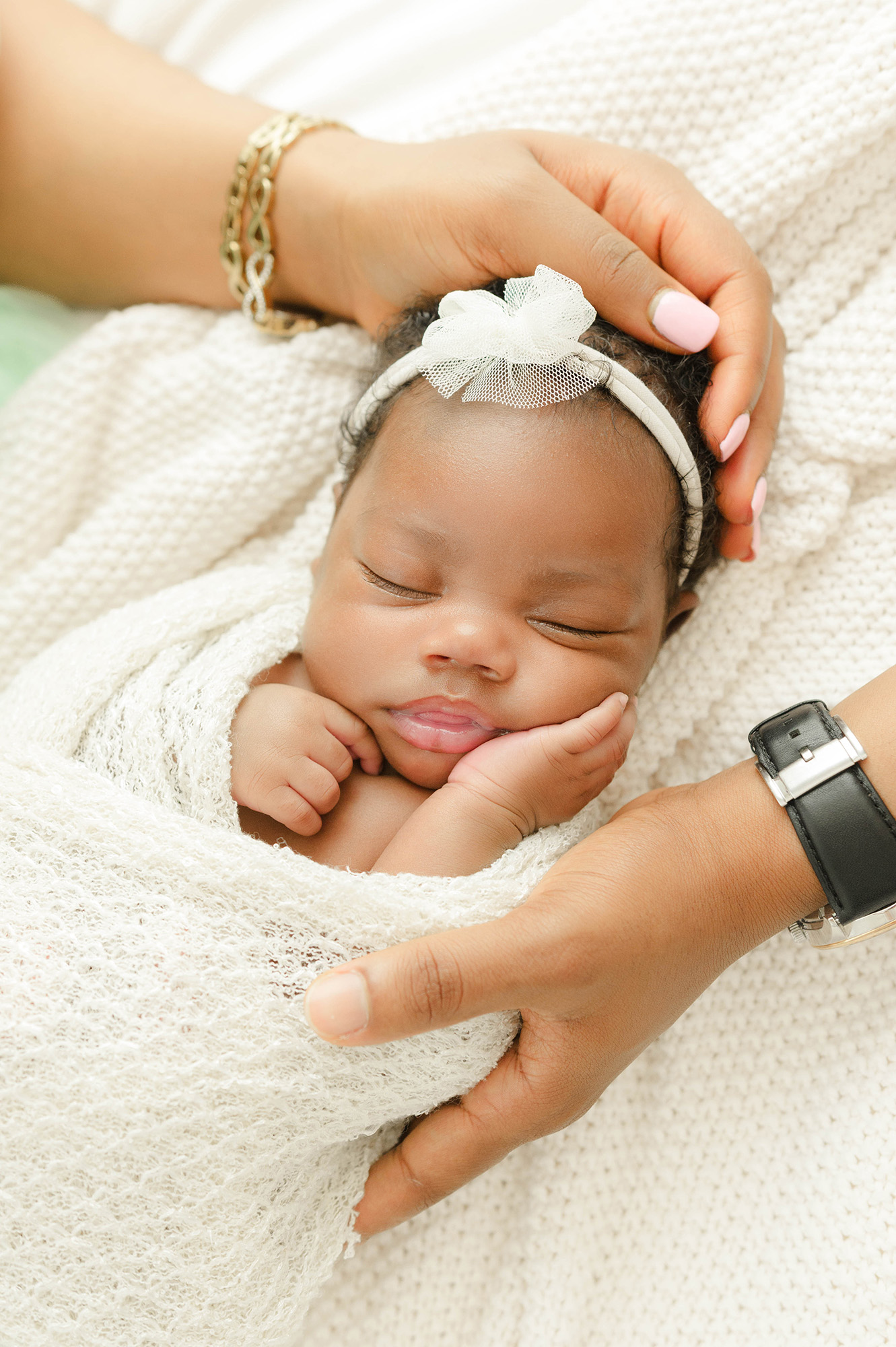 A newborn baby sleeps with a hand on her cheek in a white swaddle while mom and dad rest hands on her head and arm