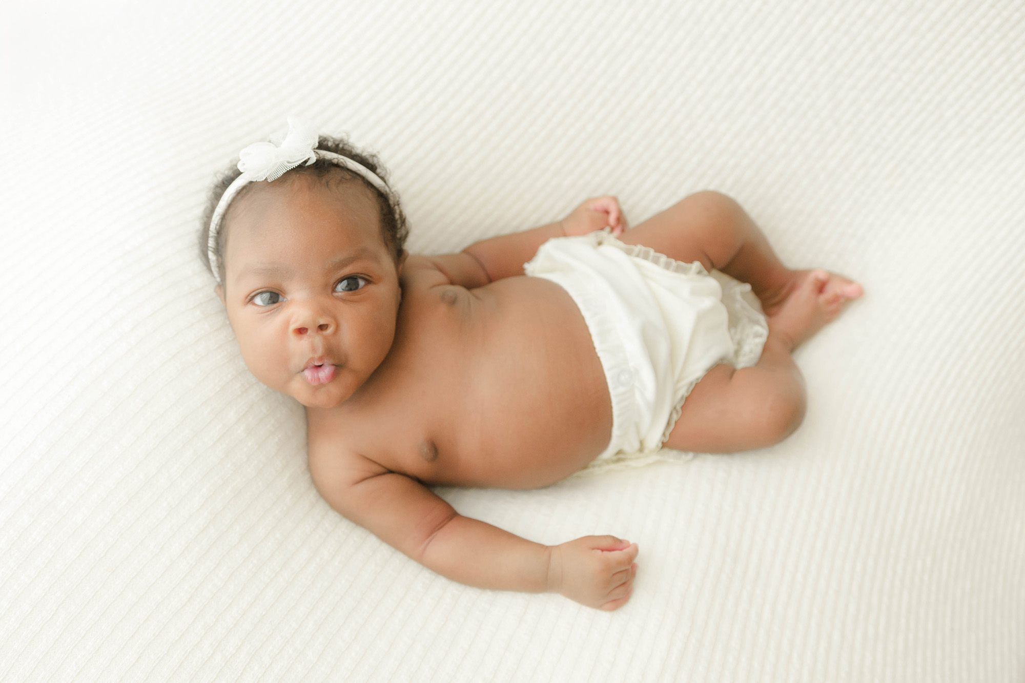 A newborn lays on a white bed in white pants with eyes open and a silly face after meeting an au pair in oklahoma city