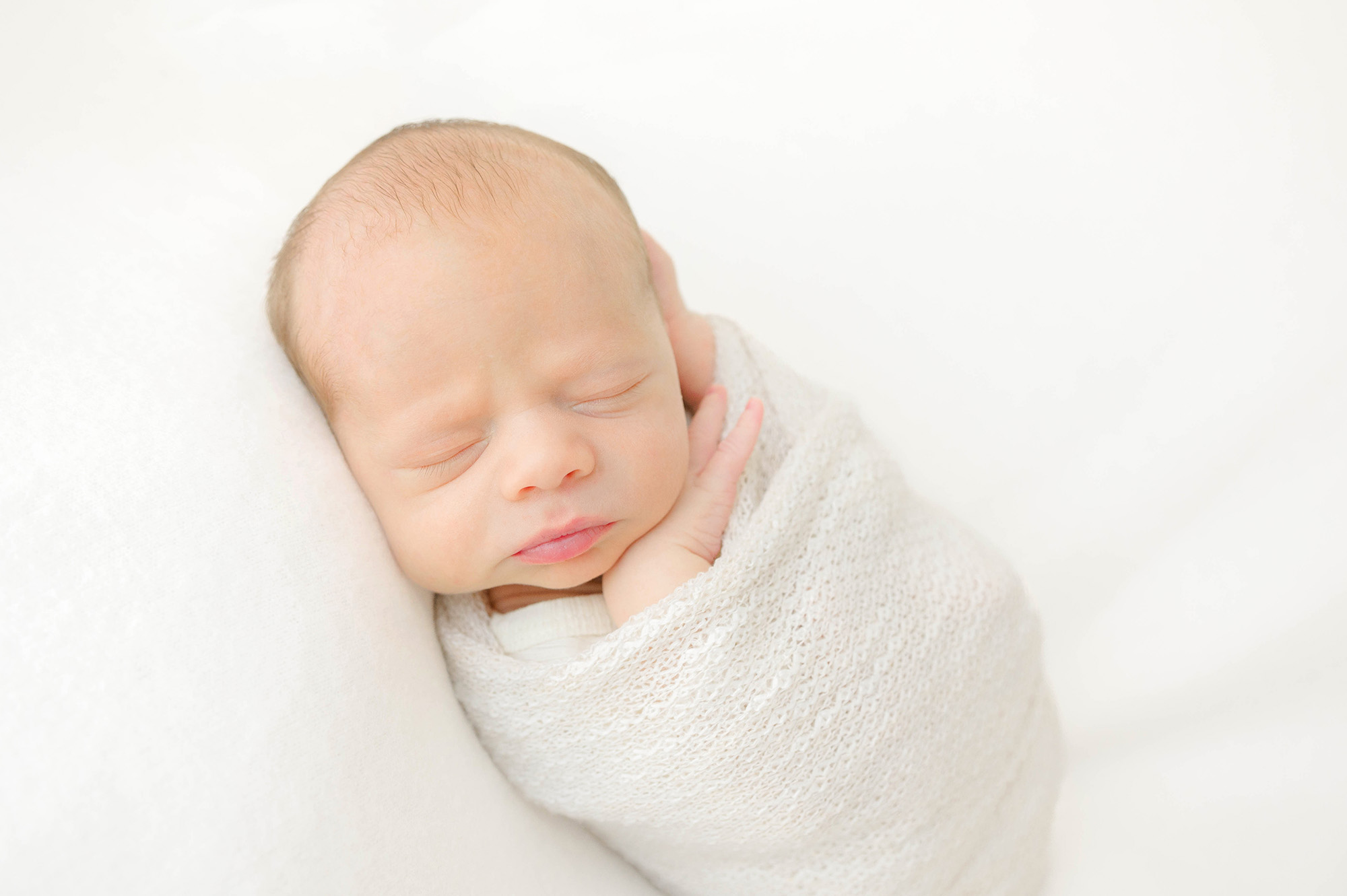 A newborn baby sleeps in a white swaddle on a white bed