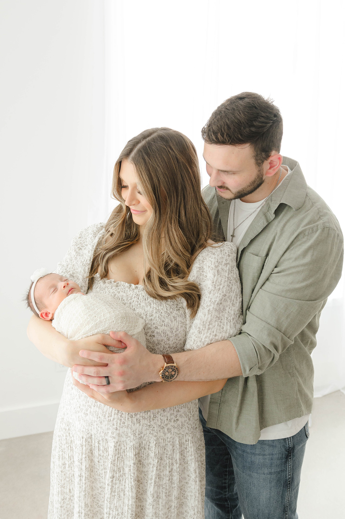 Happy new parents stand in a studio cradling their sleeping newborn daughter after meeting babysitters in okc